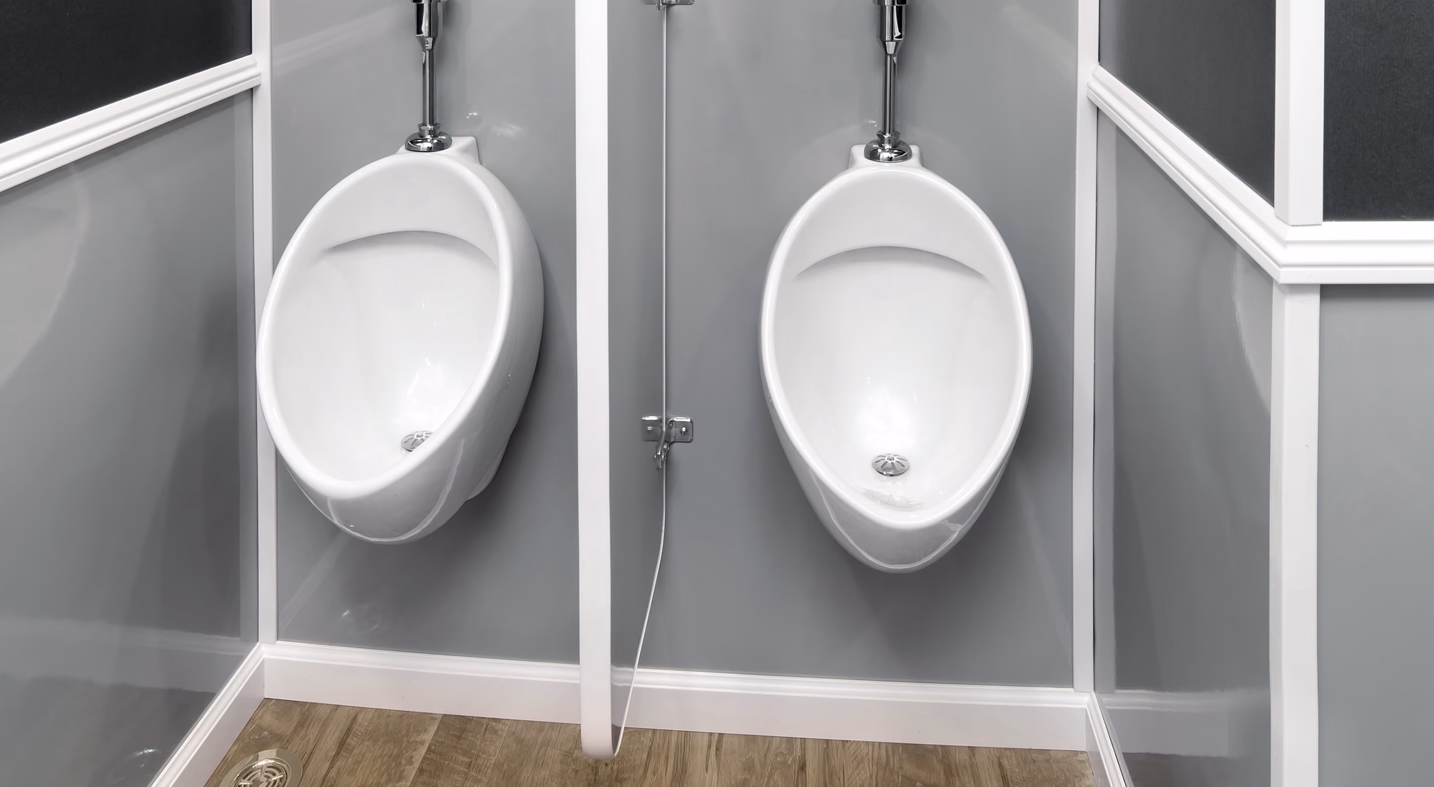 Two white urinals mounted on a grey wall, separated by a white partition. Wood-look flooring