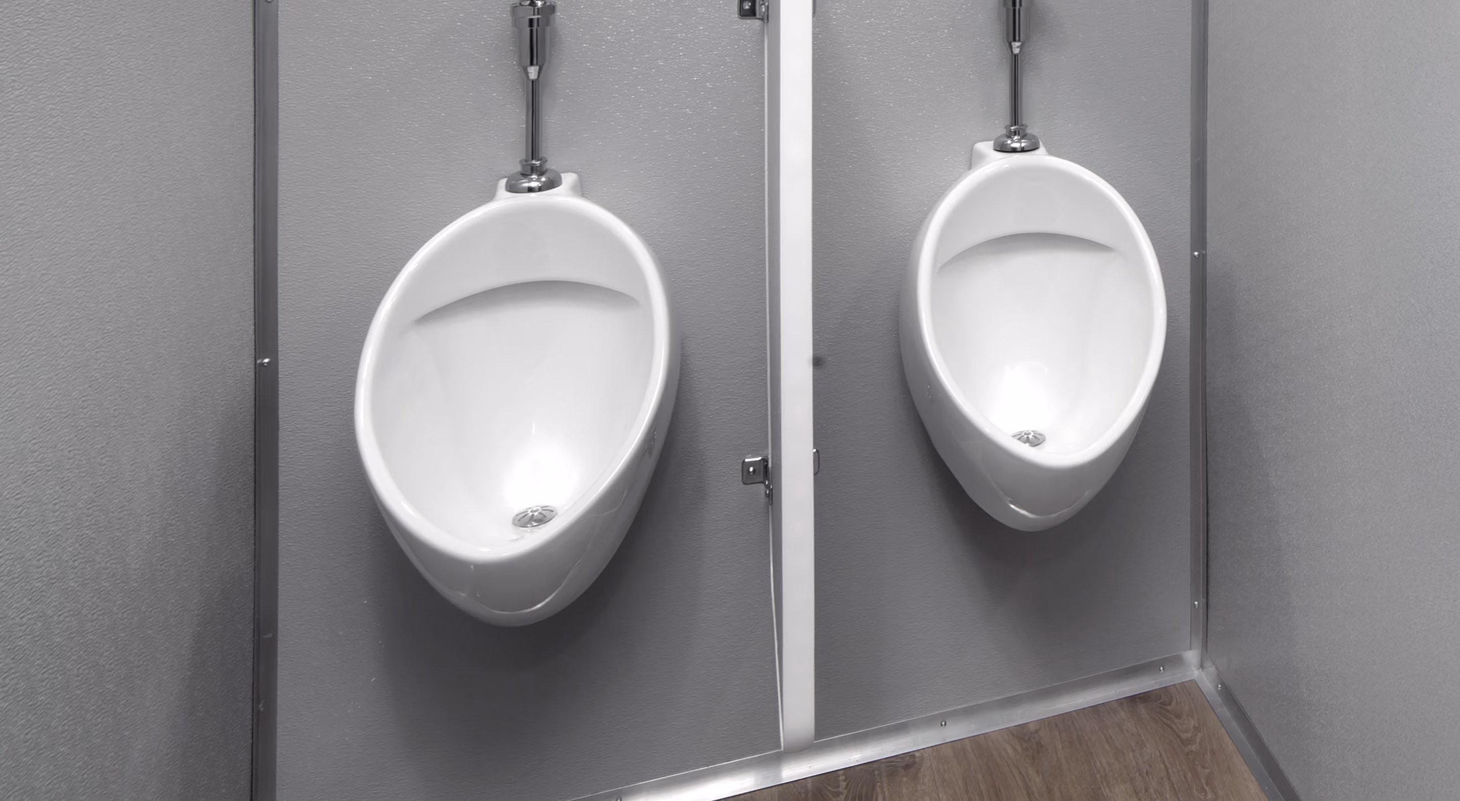 Two white urinals mounted on gray walls, separated by a metal partition. Wood laminate flooring