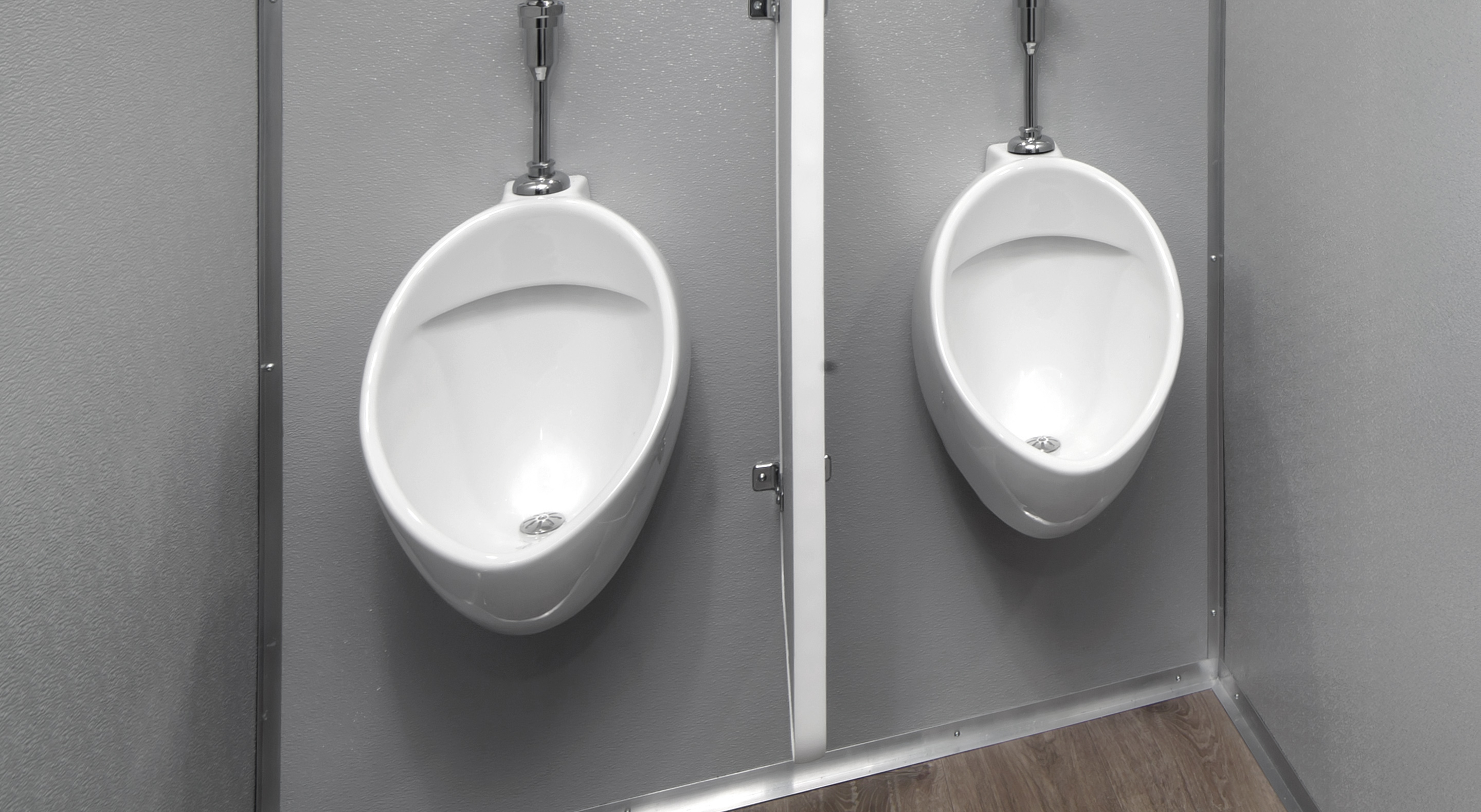 Two white urinals mounted on gray walls, separated by a metal partition. Wood laminate flooring