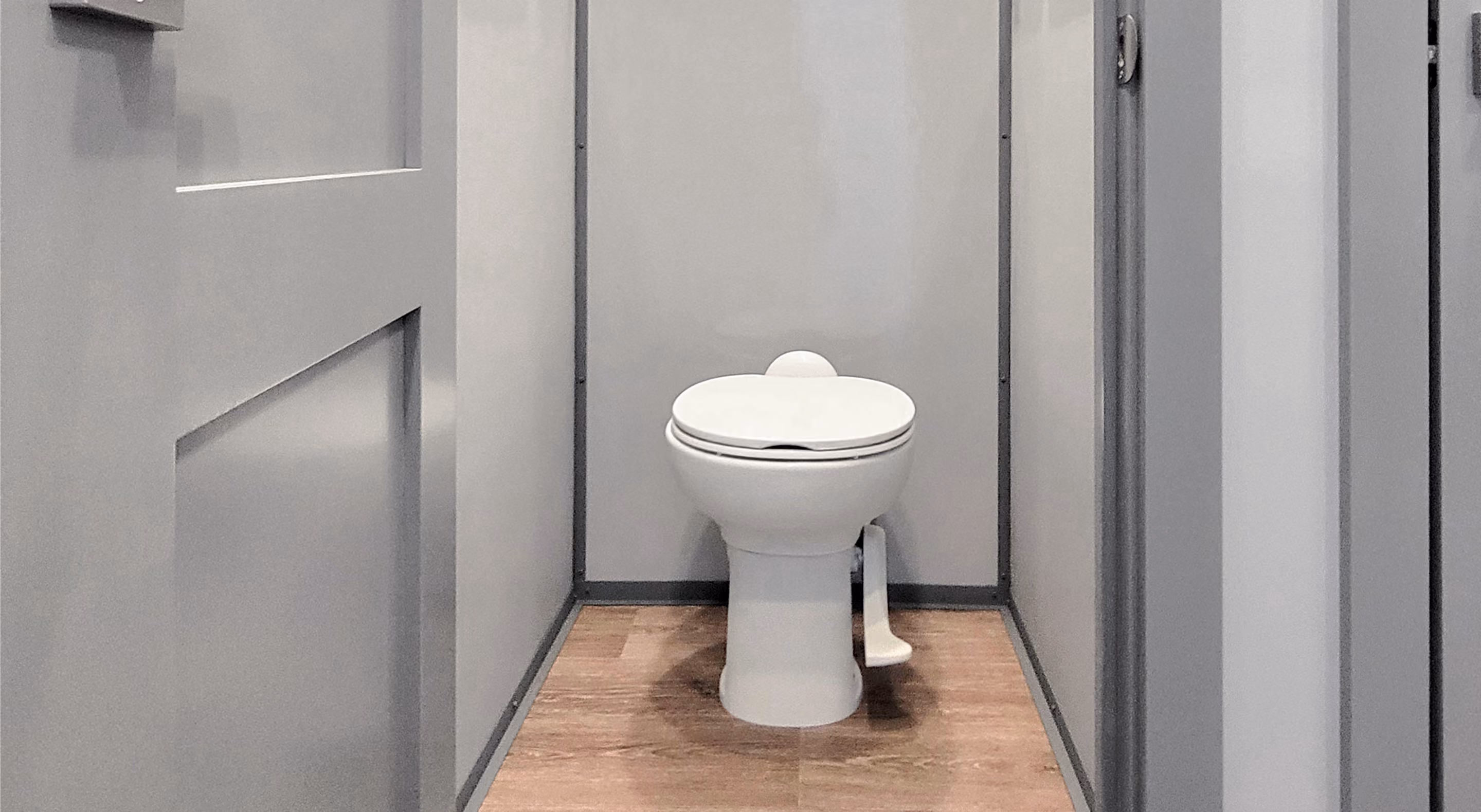 White toilet with closed lid inside a small gray restroom stall with wood floor.