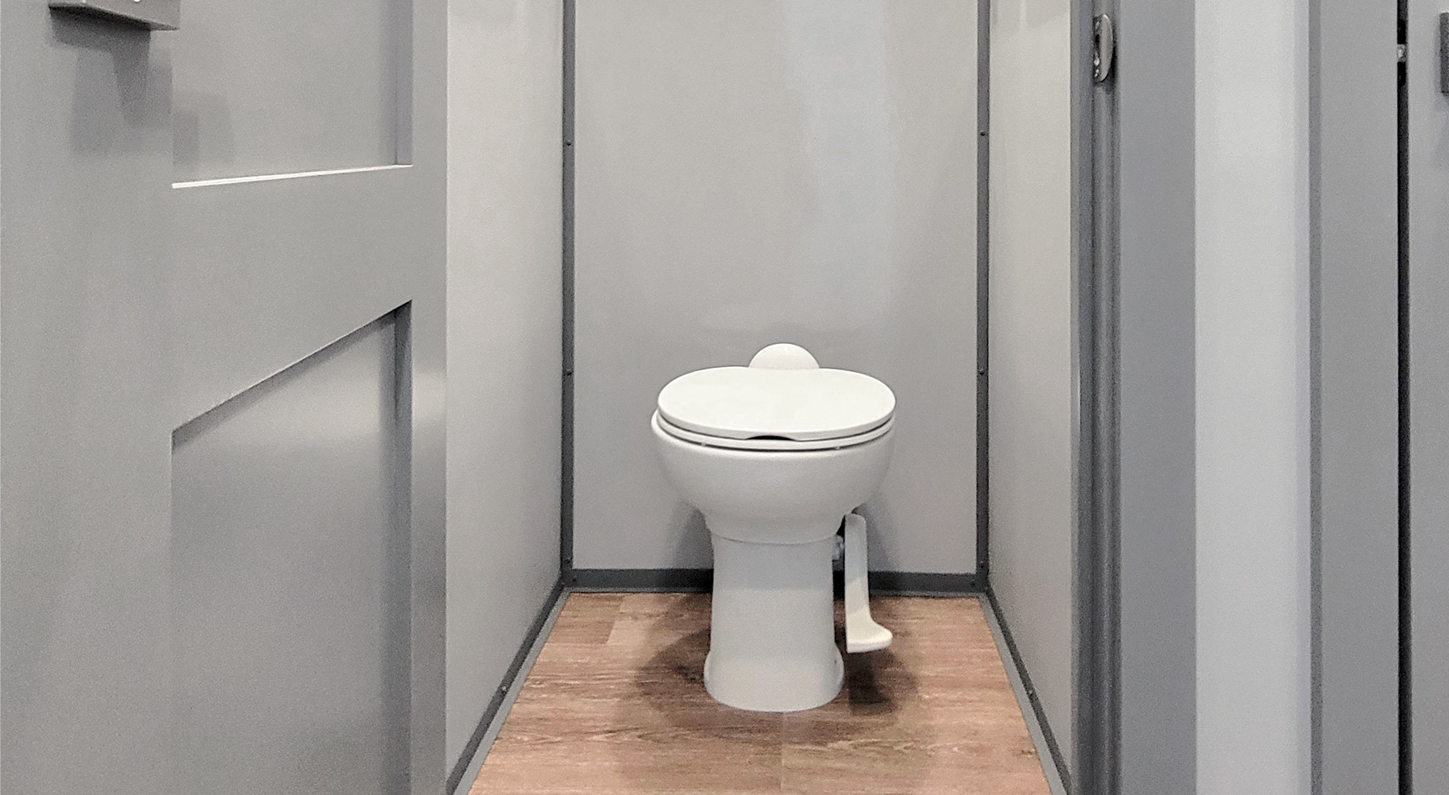 White toilet with closed lid inside a small gray restroom stall with wood floor.