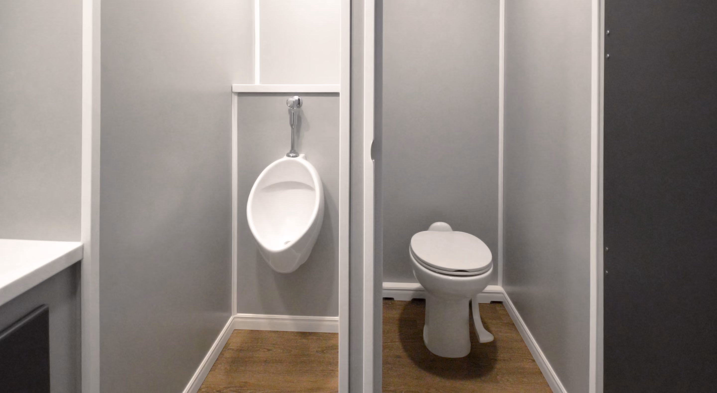 Two adjacent bathroom stalls, one with a urinal and the other with a closed toilet on wood flooring.
