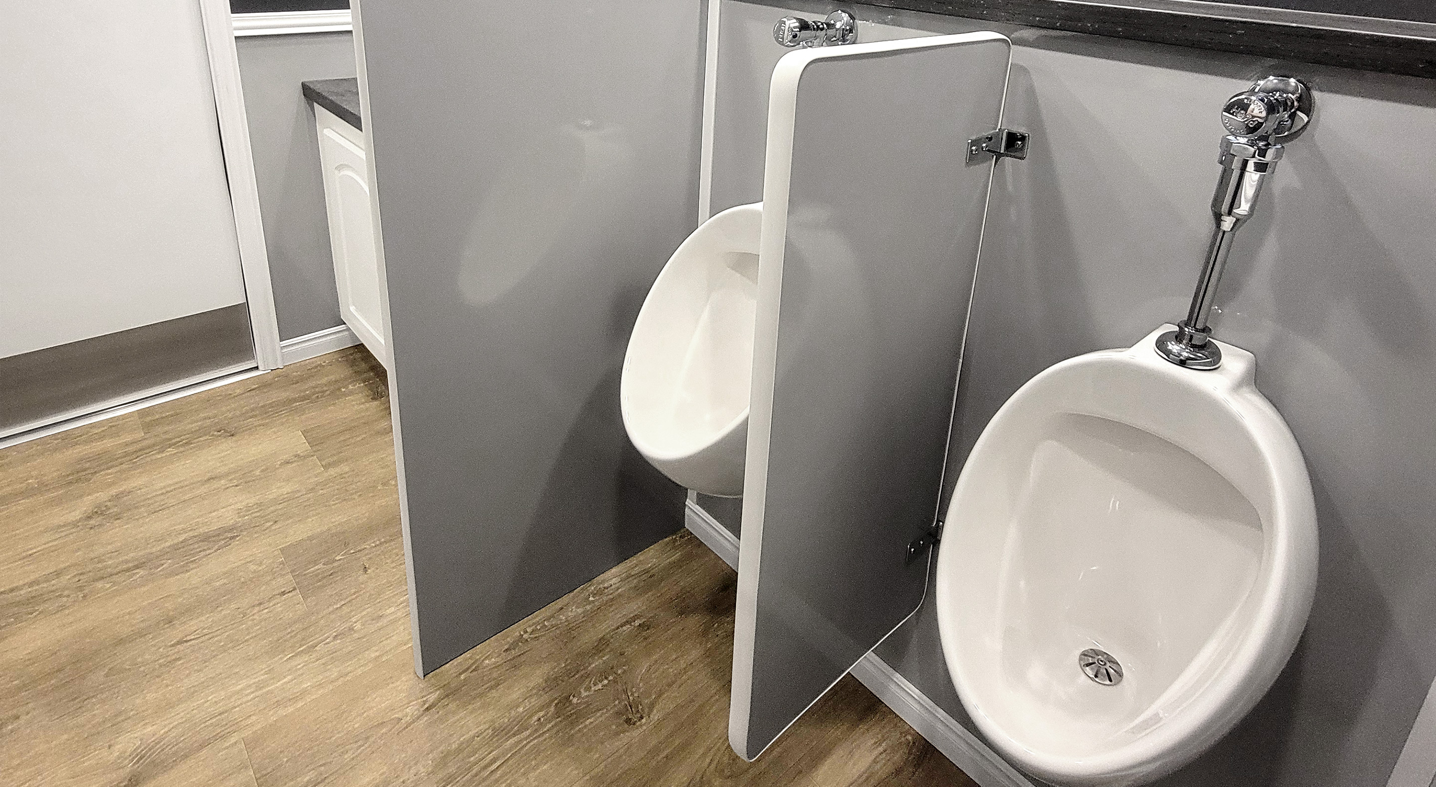 Two white urinals with a gray privacy divider between them on a wood floor against gray wall.