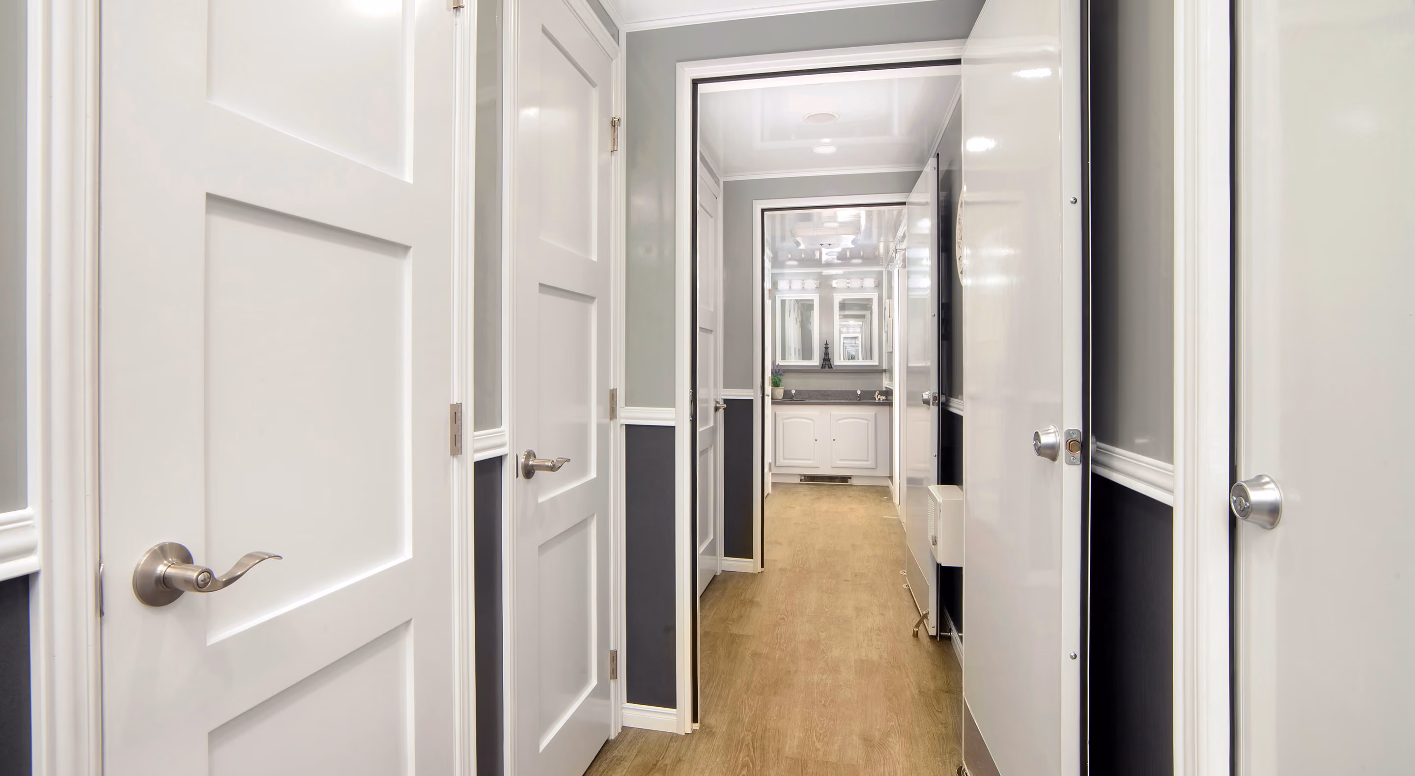 A hallway with white doors leading to a well-lit bathroom with mirrors and modern fixtures.