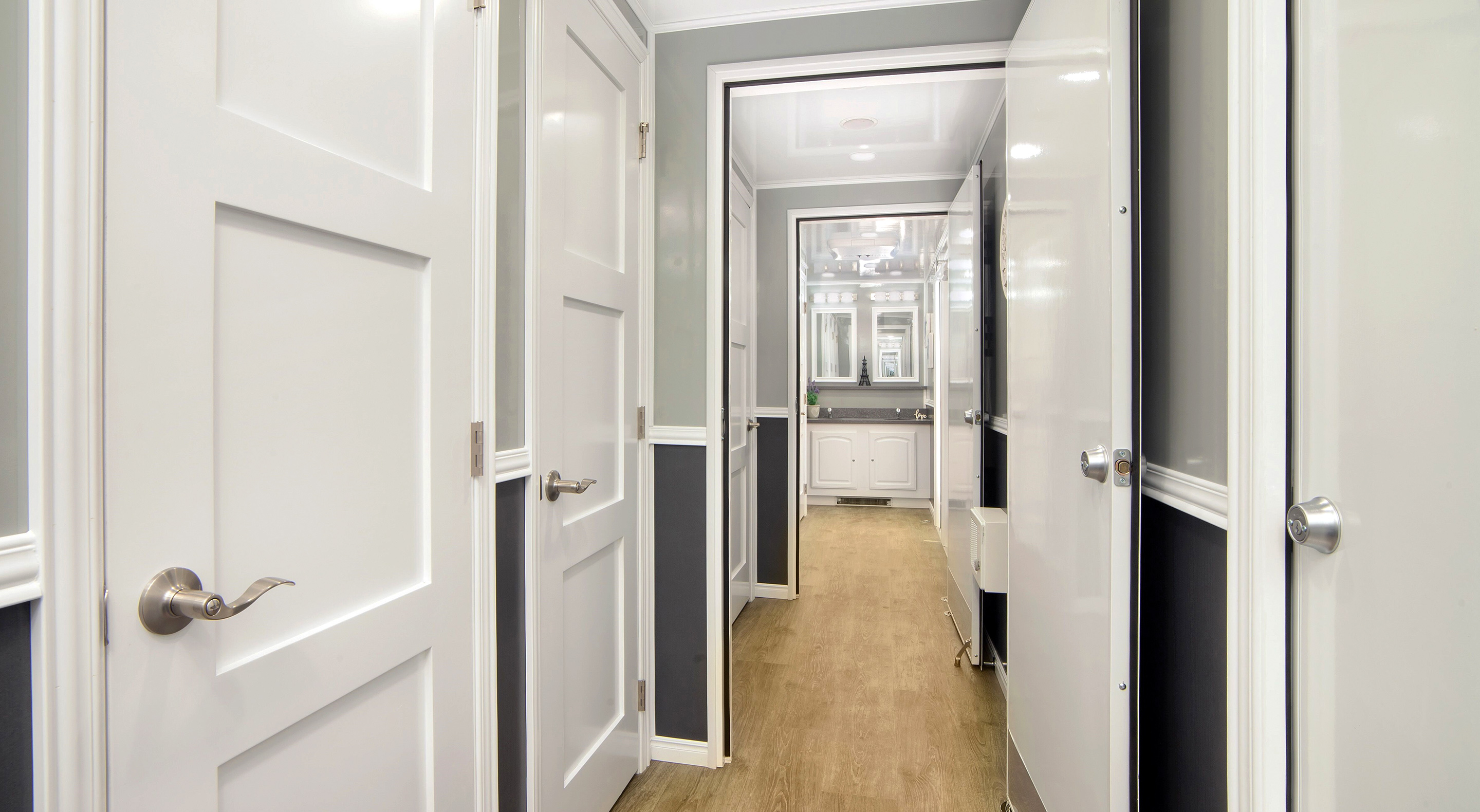A hallway with white doors leading to a well-lit bathroom with mirrors and modern fixtures.