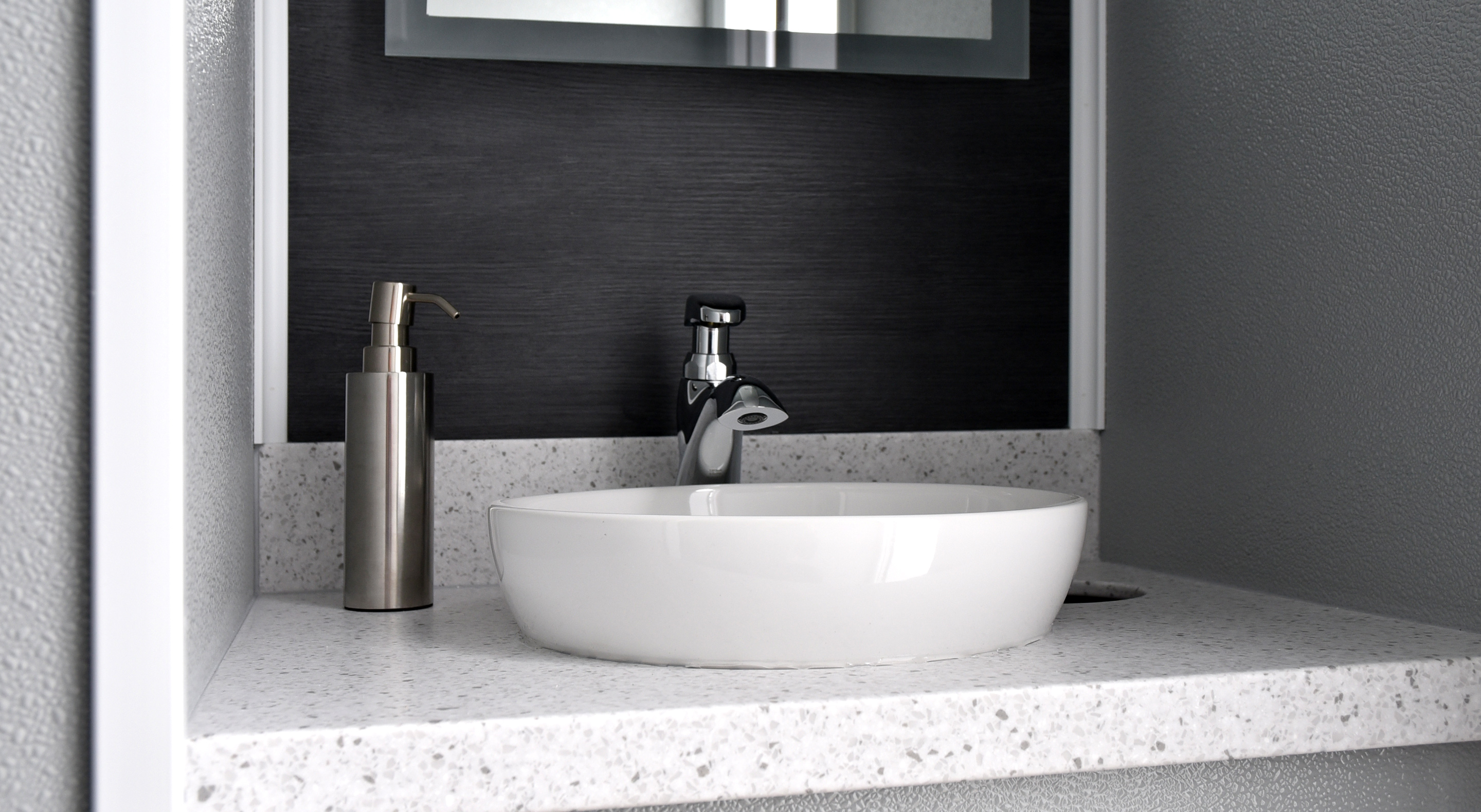 White oval sink with chrome faucet and stainless steel soap dispenser on speckled countertop.