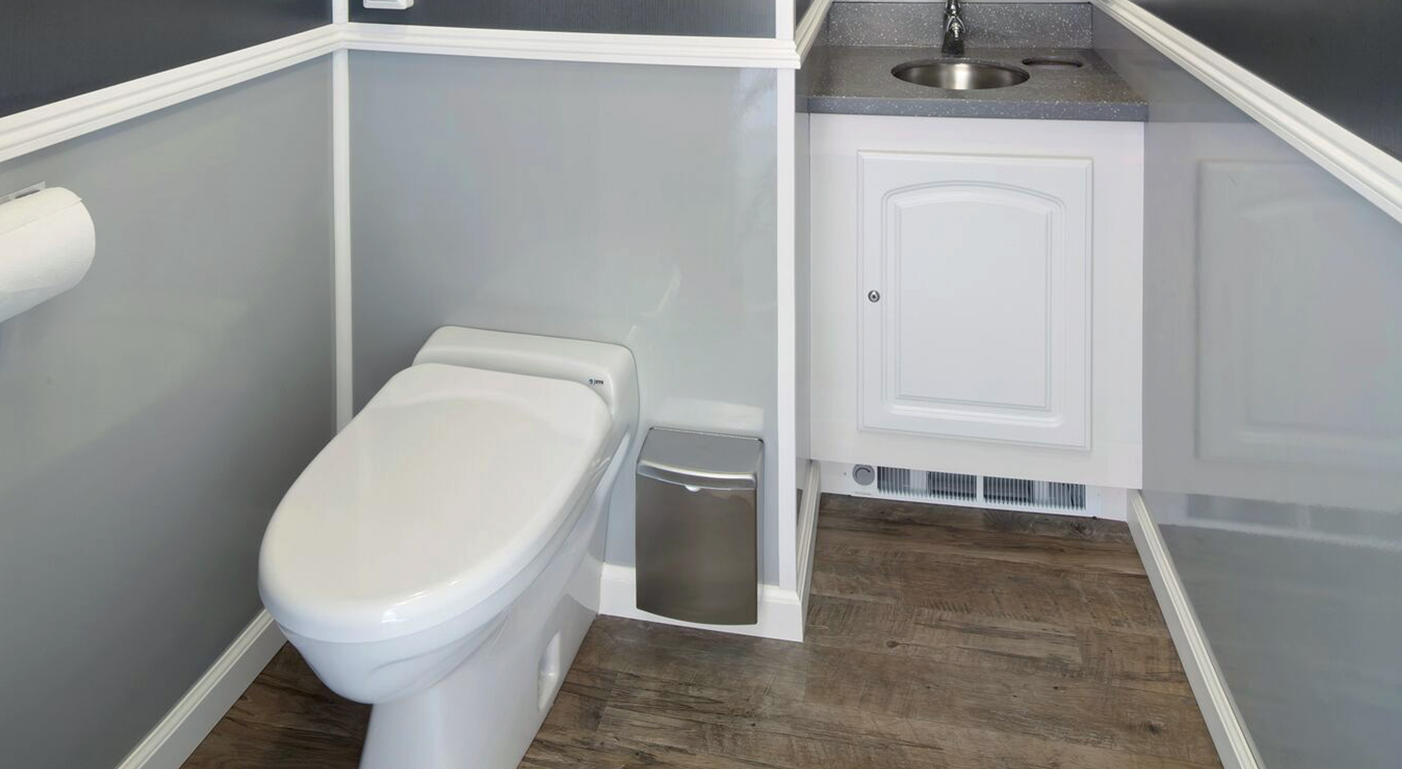 Small bathroom with white toilet, metal trash bin, gray walls, wood floor, and white sink cabinet.