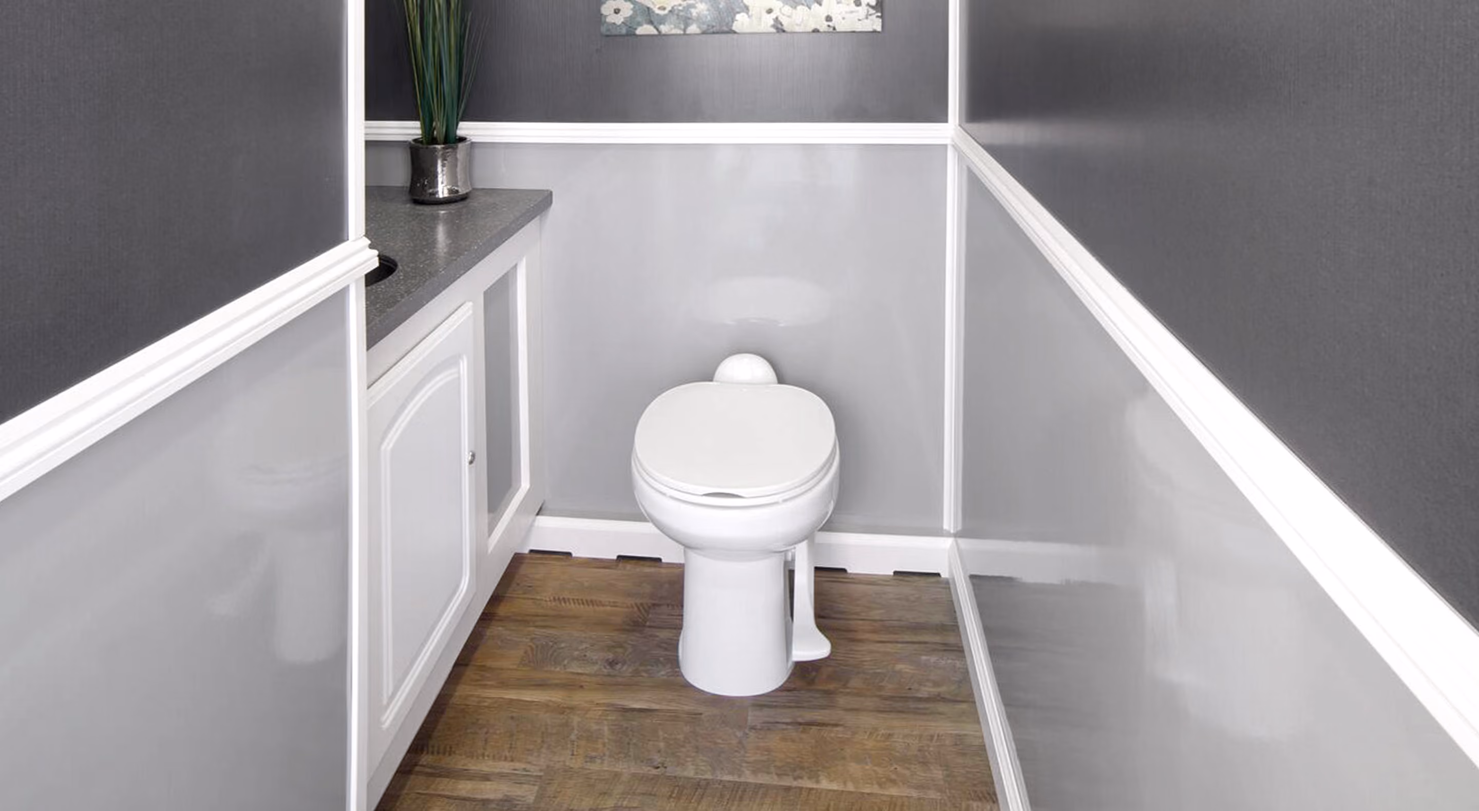 Small modern restroom with white toilet, wood floor, gray walls, and white cabinetry with plant.