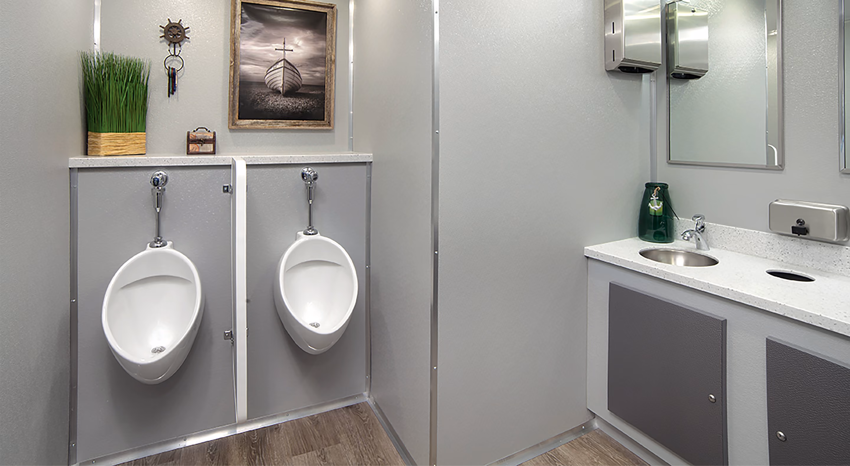 Modern restroom with two urinals, a sink, a mirror, green vase, grass plant, and framed boat photo.