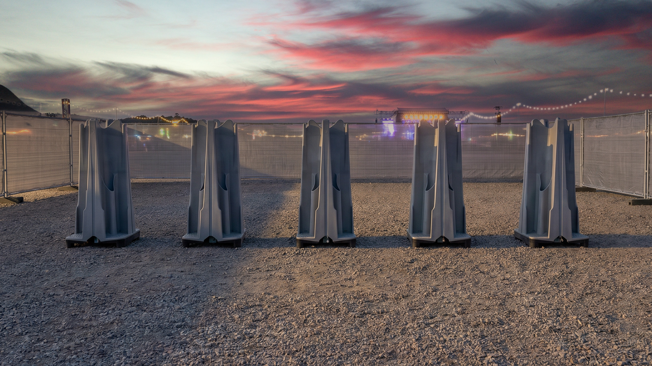 Five large, gray plastic containers stand in a row on gravel under a colorful sky.