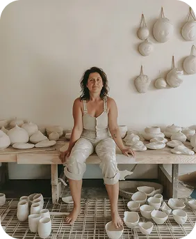 Woman sitting barefoot on a wooden table surrounded by handmade ceramic pottery including bowls and vases in a minimalist studio.