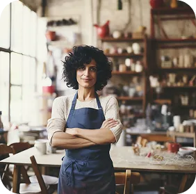 Confident woman with curly hair wearing a blue apron in a cozy, well-lit kitchen or cafe.
