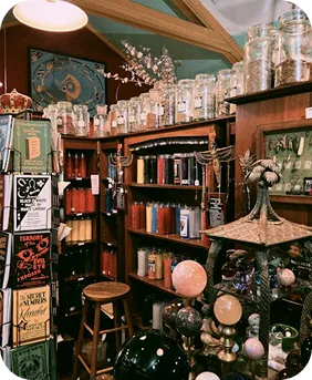 Interior of a vintage apothecary shop with shelves of glass jars, bottles, candles, and mystical decor including globe-like objects and a wooden stool.