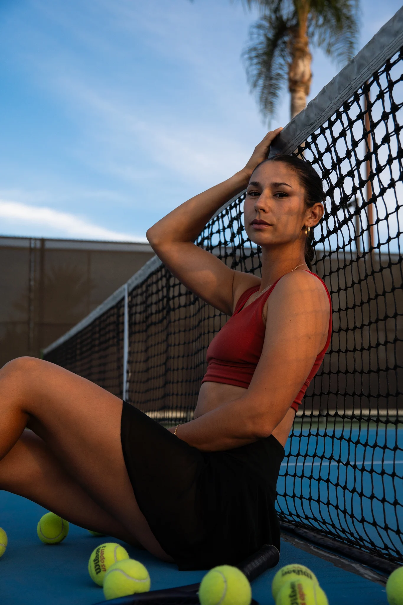 A female athlete leaning against a tennis net wearing a black skirt and a red sports bra.
