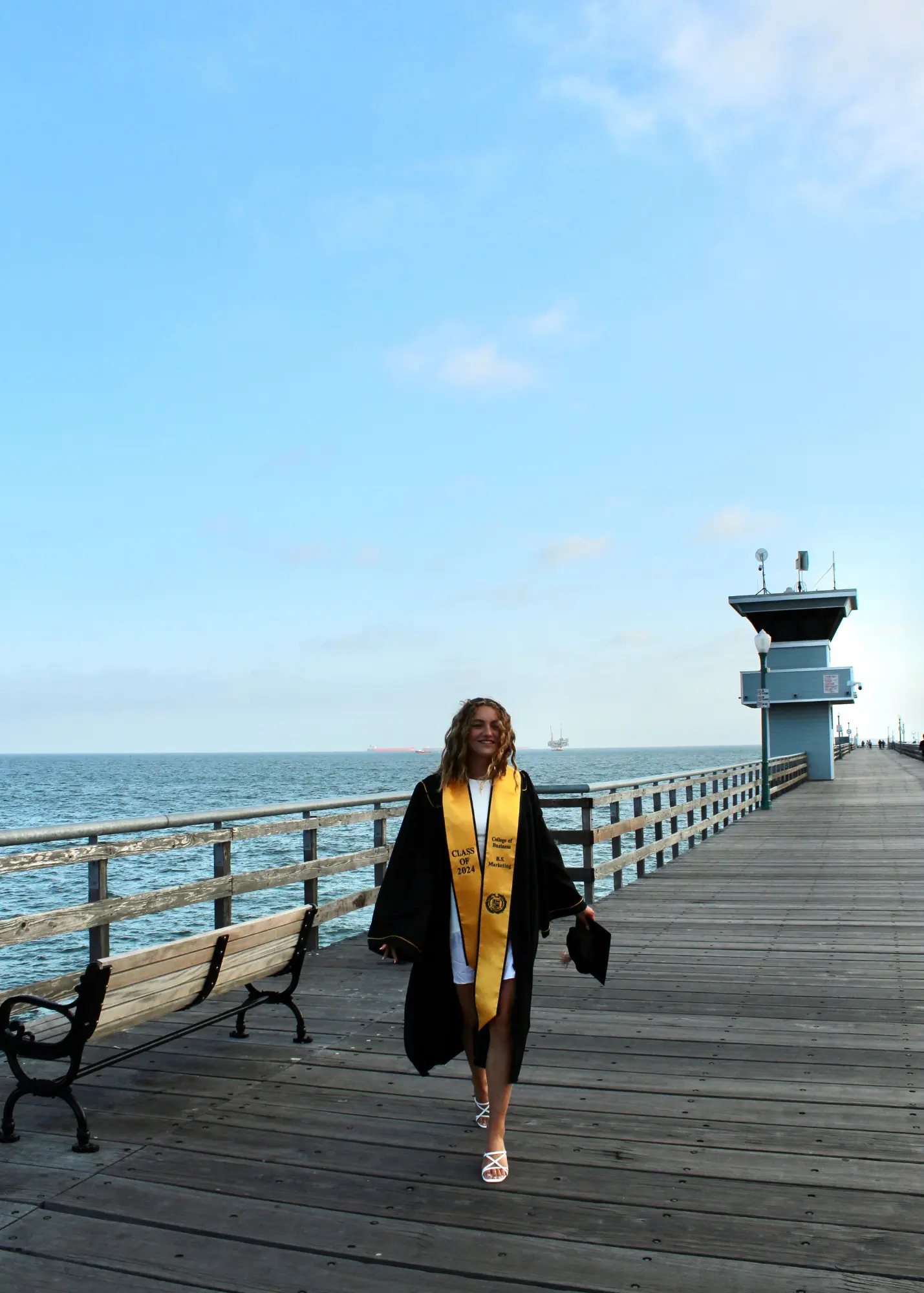 A recent female graduate walking along a beach pier, wearing a white dress, black coat and cap, with a yellow sash.