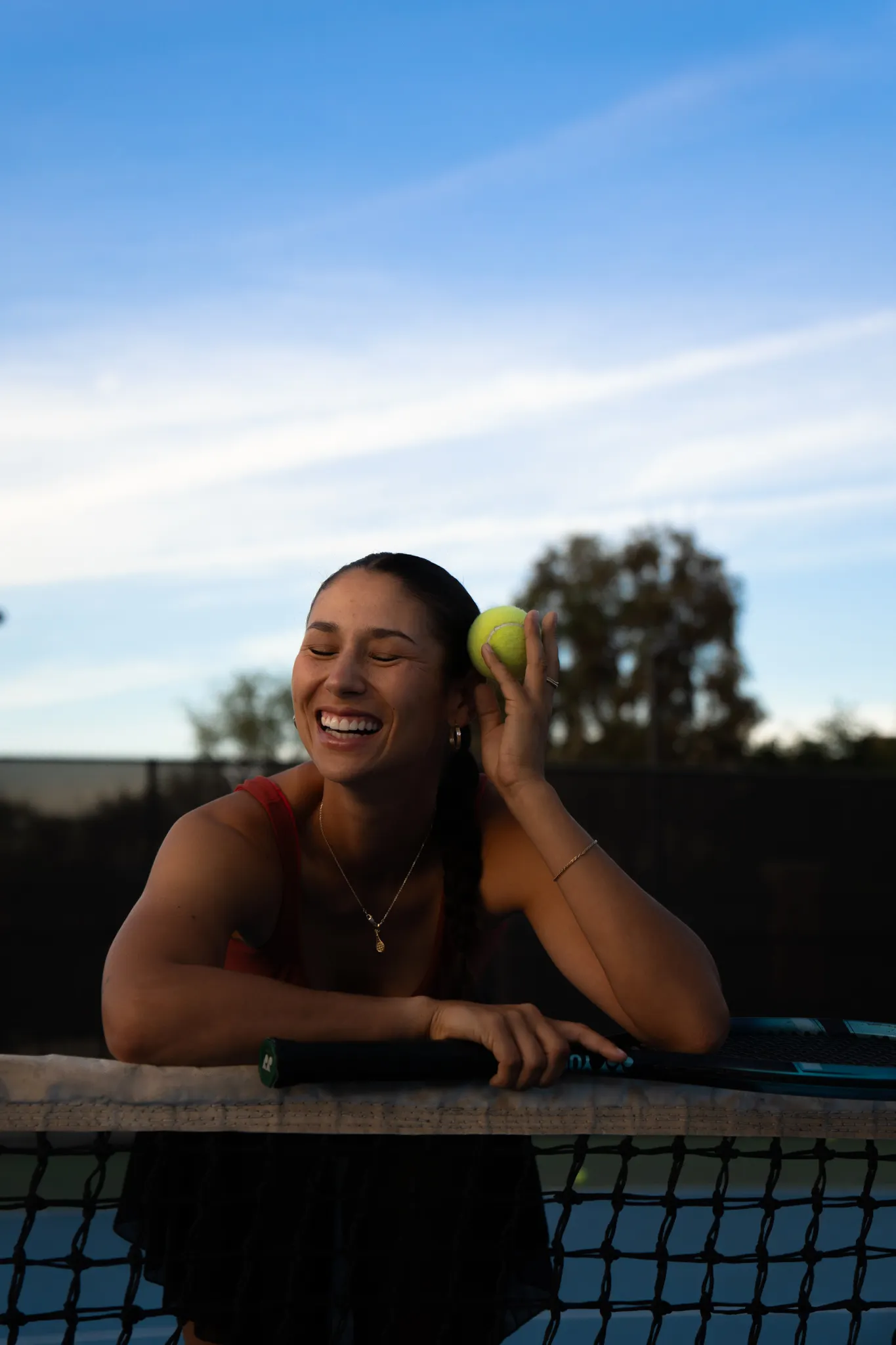 A girl smiling, leaning against a tennis net.
