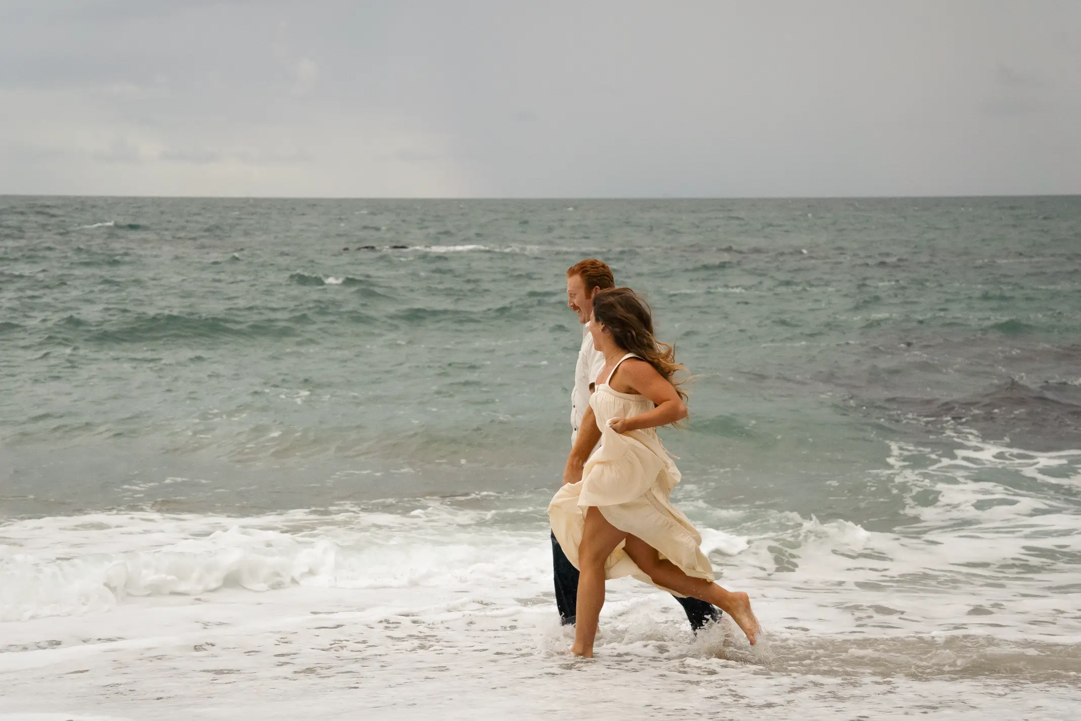 A newly engaged couple running across a beachy shore.