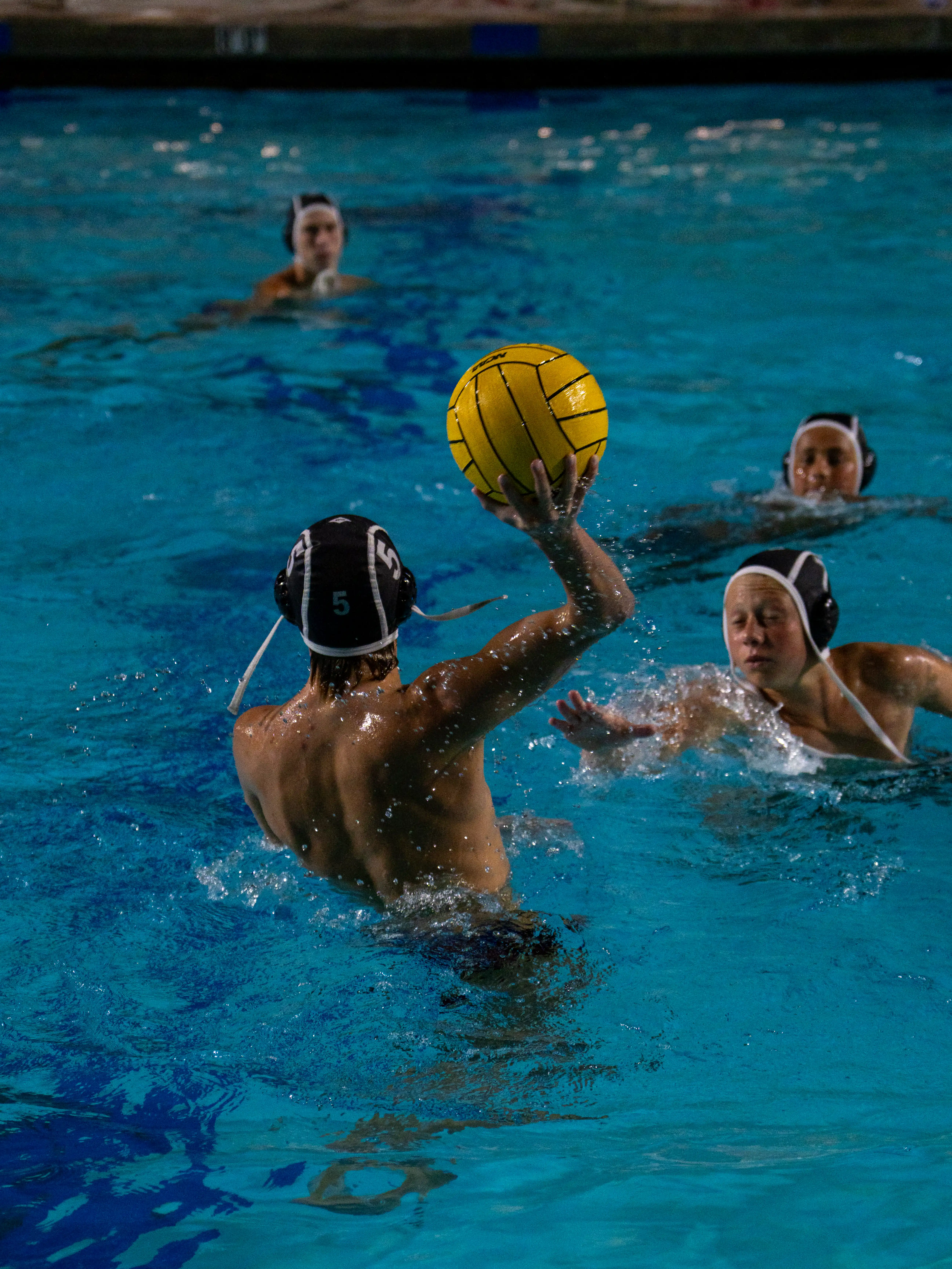 Water polo player wearing cap number 5 prepares to throw a yellow ball to his teammates in the pool.