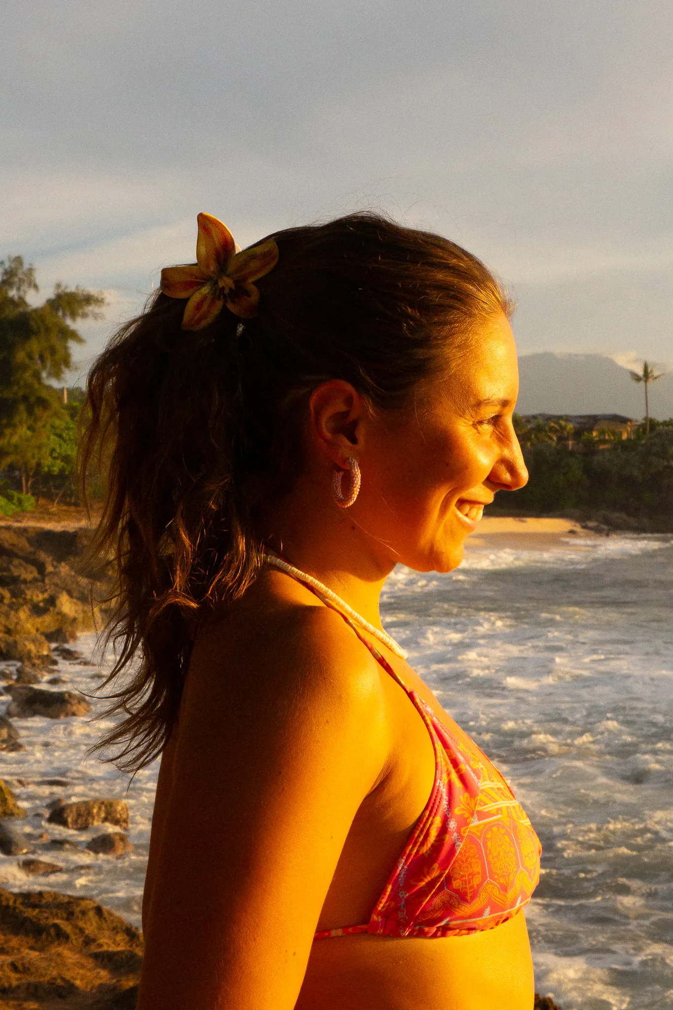 Smiling woman with a flower in her hair wearing a pink bikini top, standing by rocky beach at sunset.