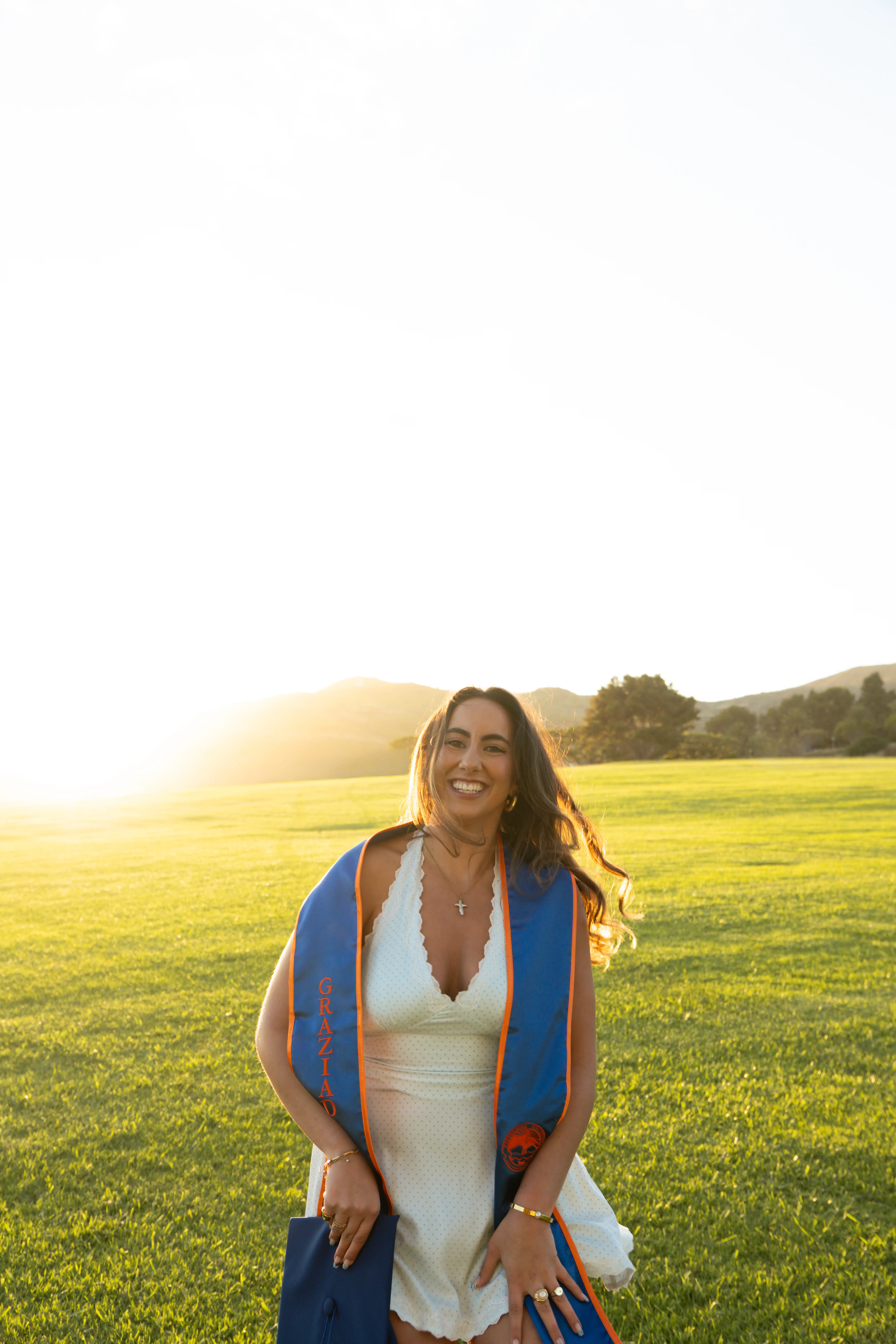 Smiling graduate in white dress wearing blue and orange stole, holding cap in sunlit green field.