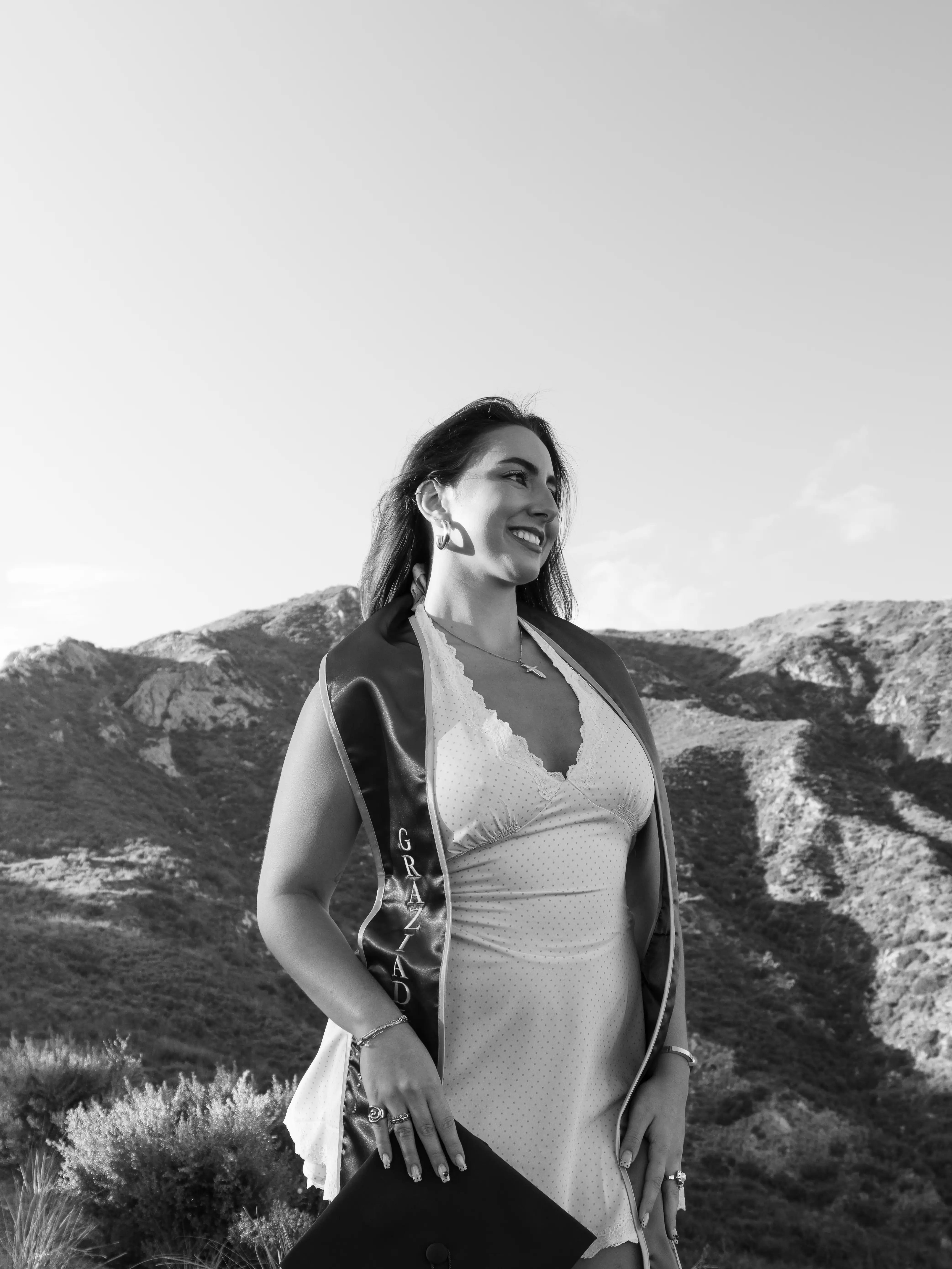 Smiling young woman in a white dress holding a graduation cap outdoors with mountains in background.