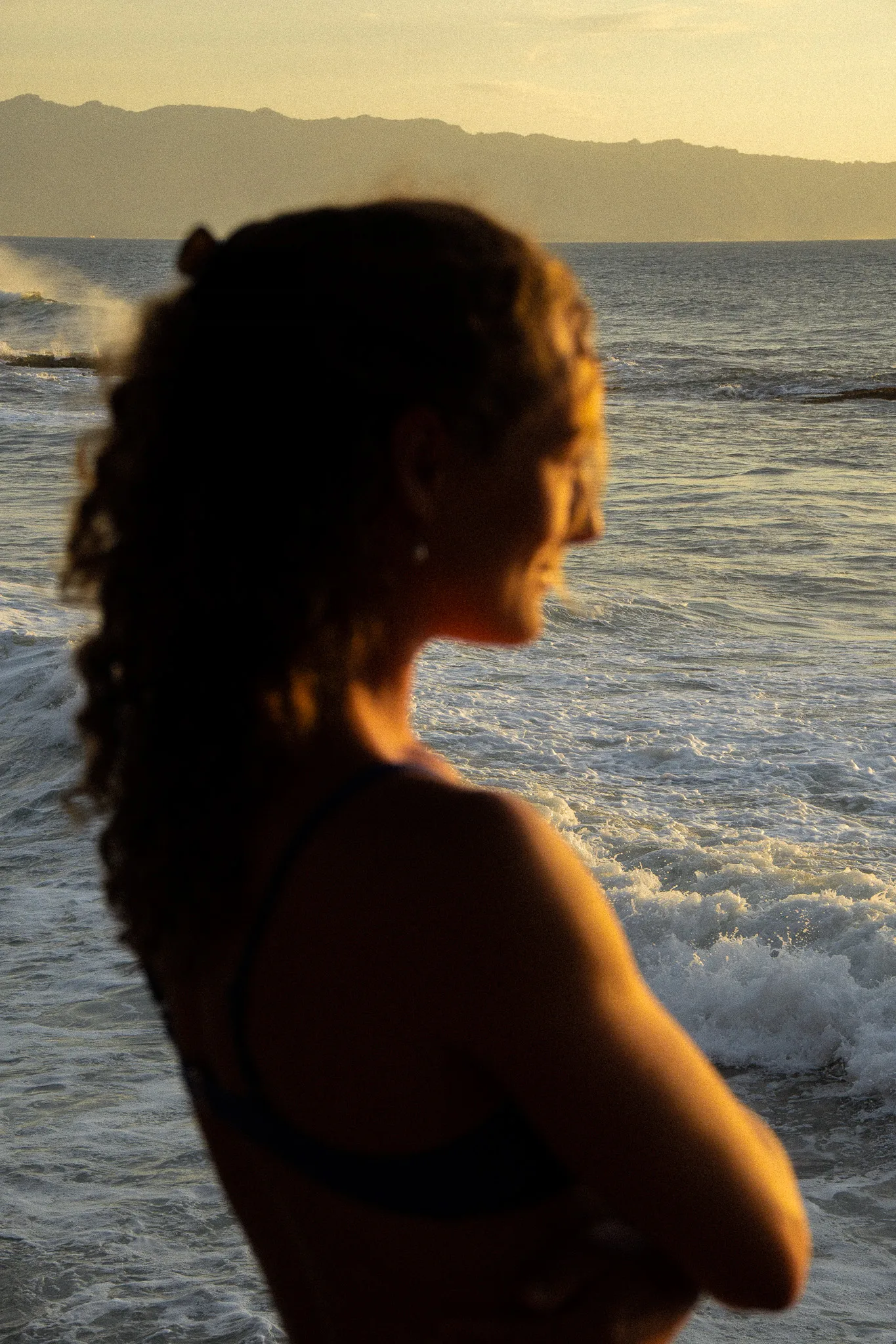 Silhouette of a woman with curly hair standing by the ocean with waves and distant mountains at sunset.