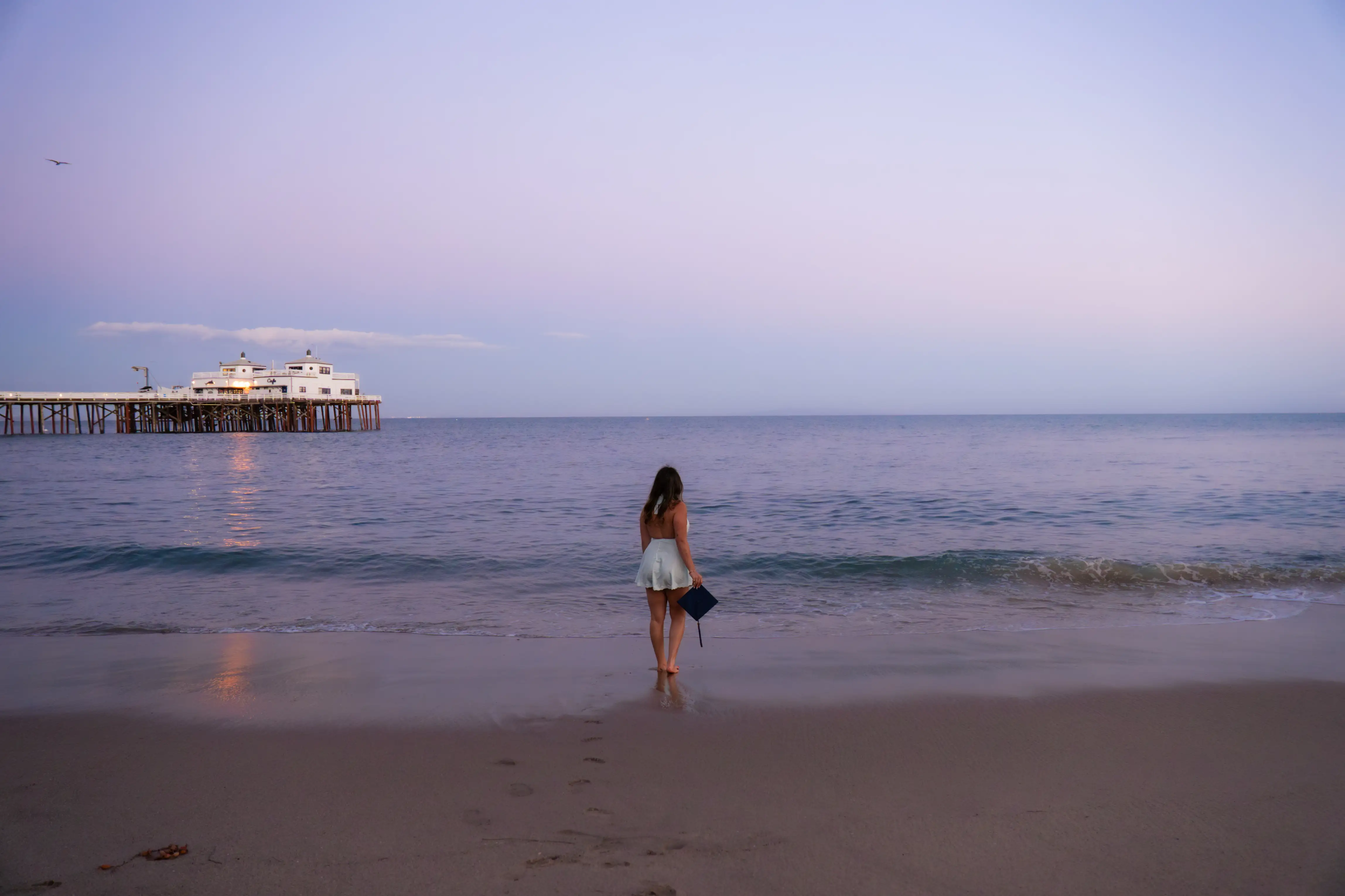 Woman in white dress holding graduation cap, standing barefoot on beach facing calm ocean at sunset.