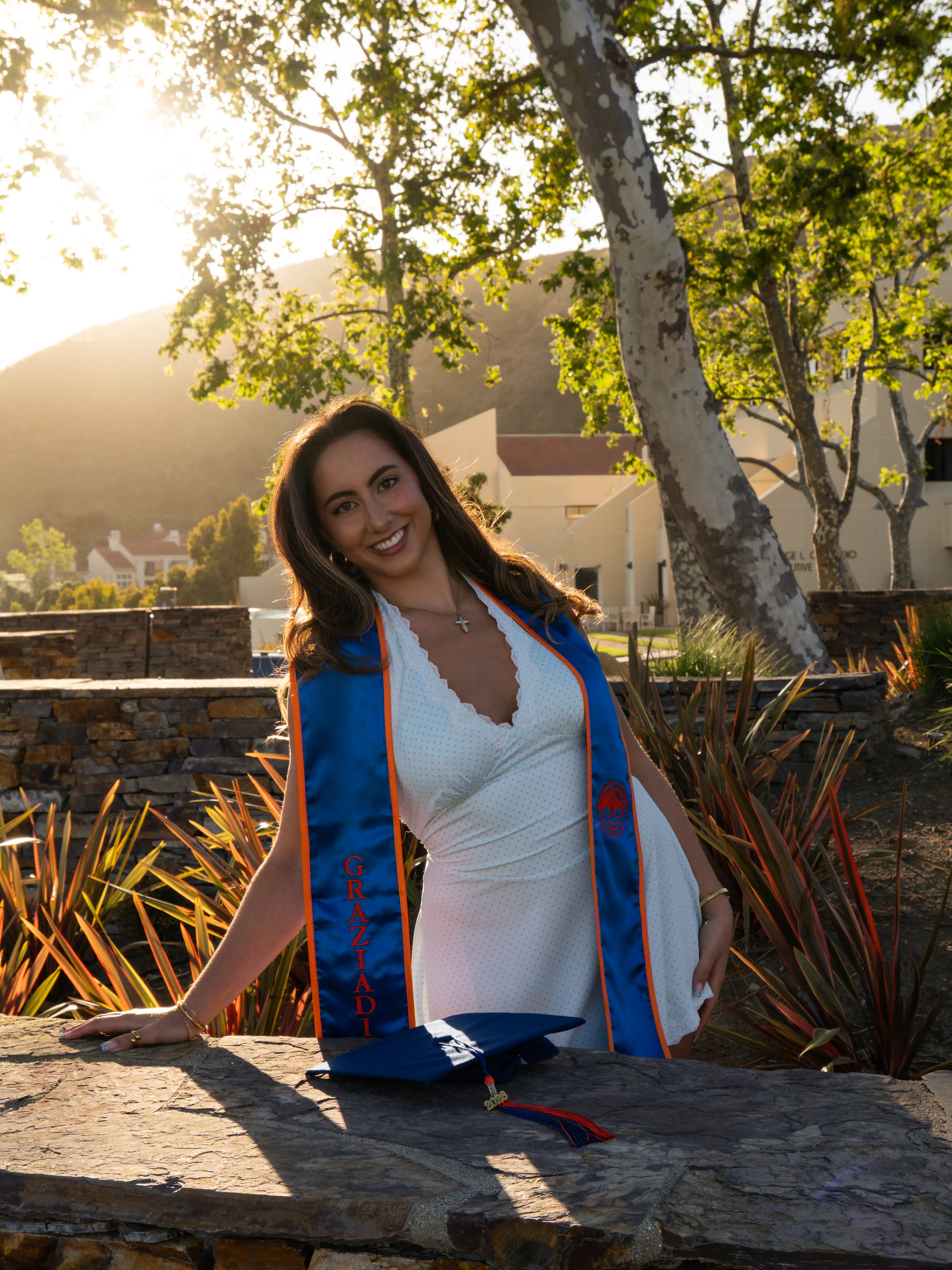 Smiling woman in white dress with blue graduation stole leaning on stone ledge with cap in sunlight.