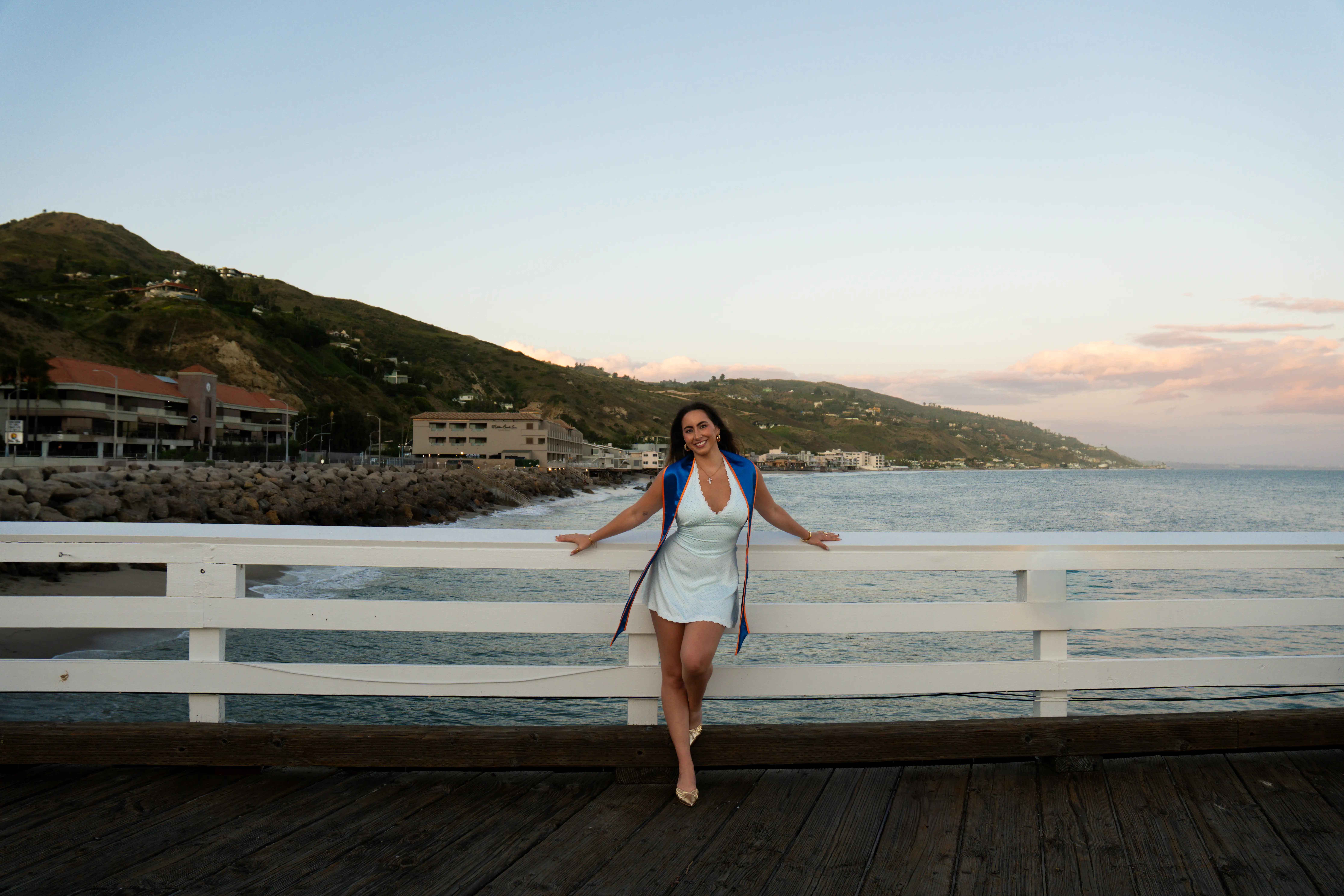 Smiling woman in white dress and blue graduation stole leaning on white railing by ocean at sunset.