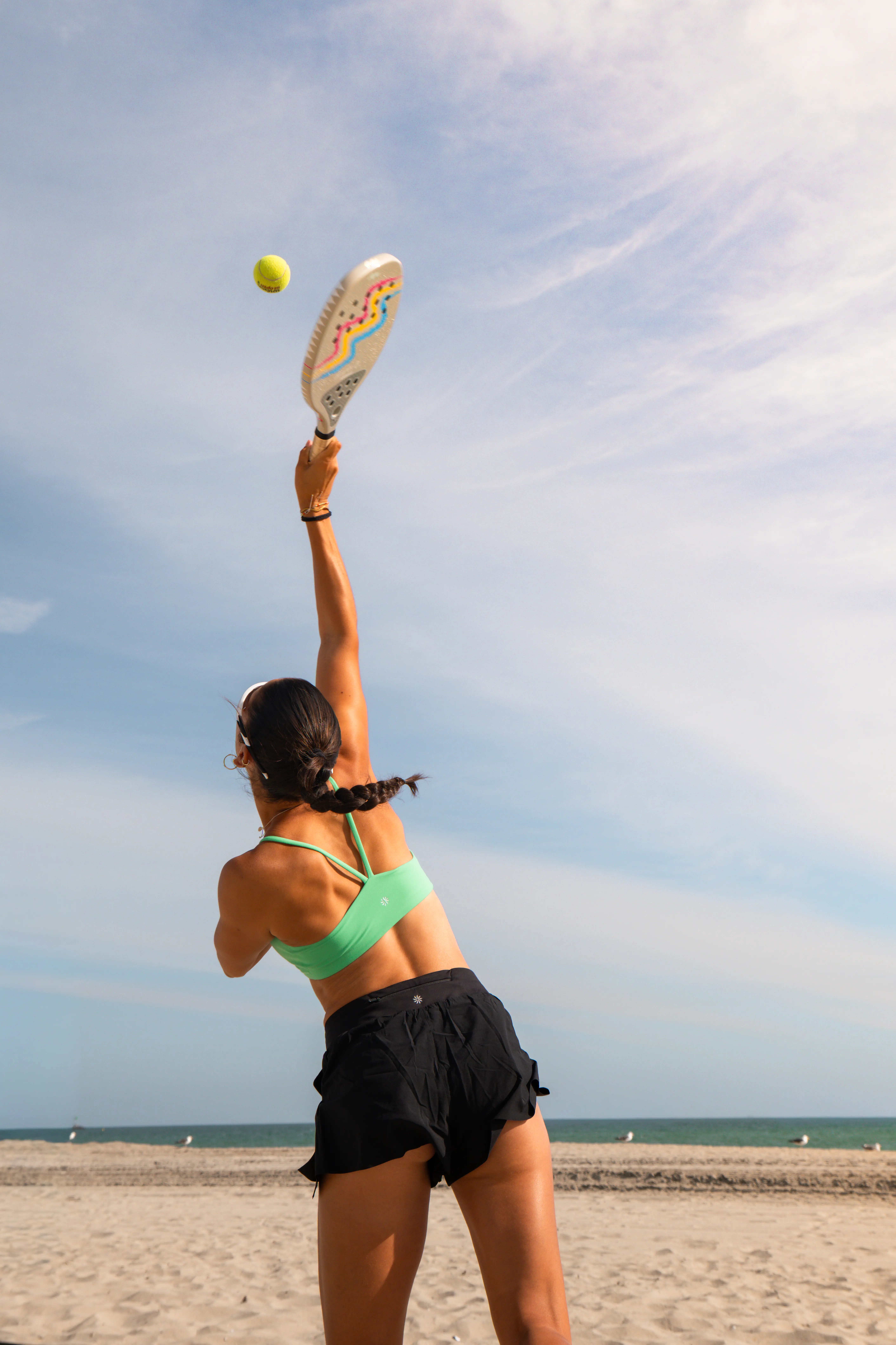 Woman in yellow sportswear raising one arm against a blue sky with wispy clouds.
