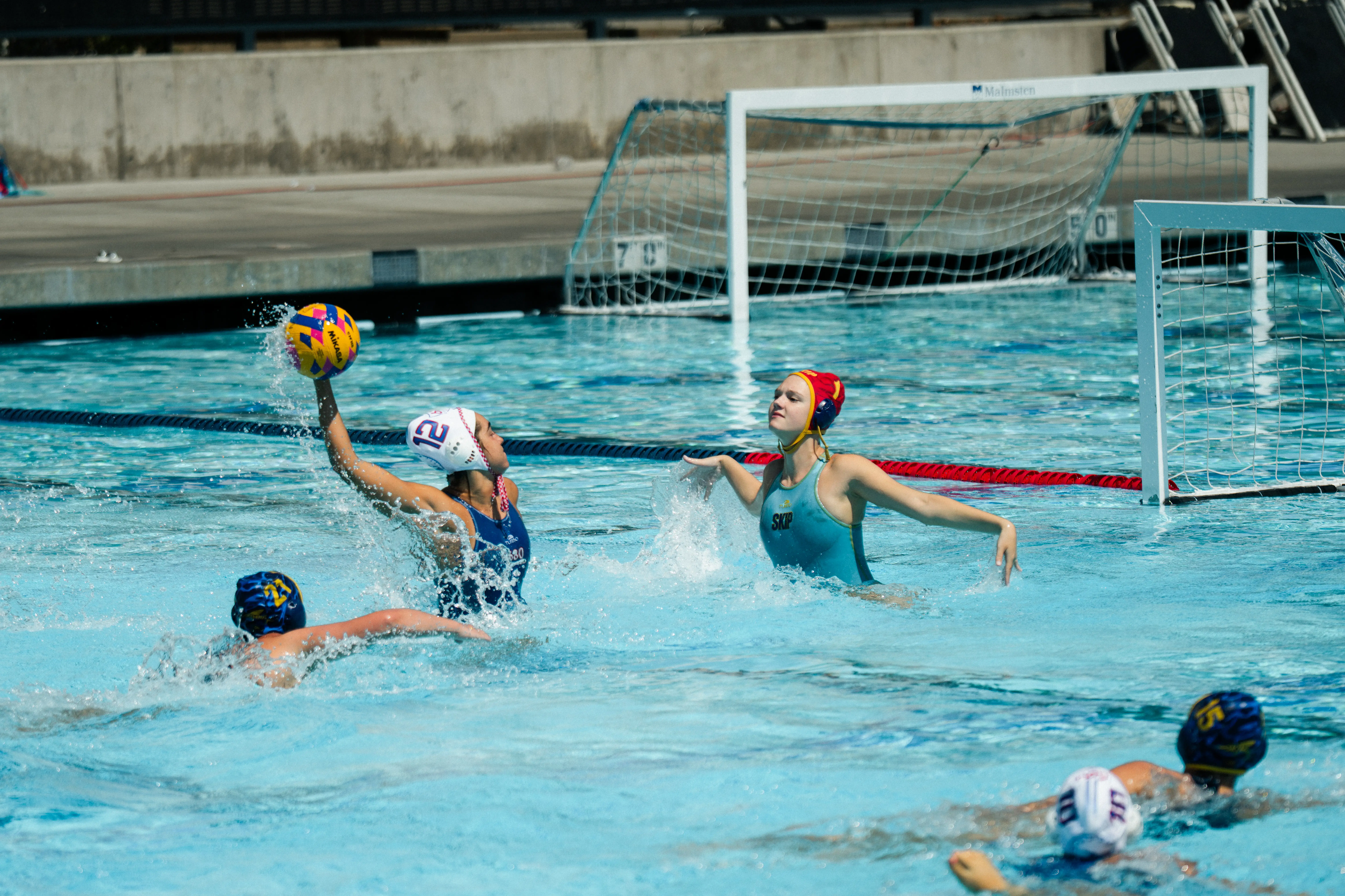 Water polo player in white cap prepares to throw ball while goalie in red cap defends goal.