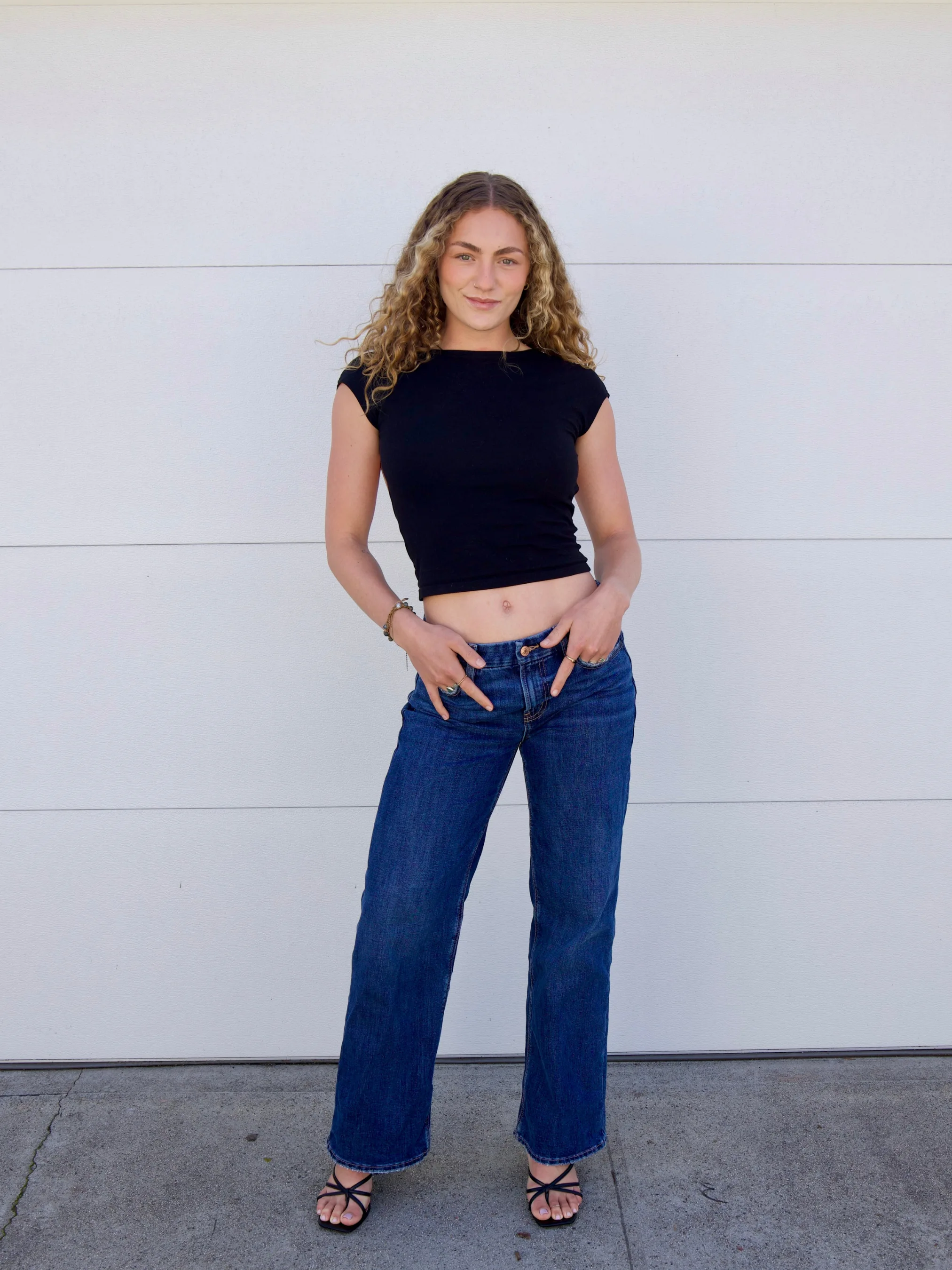 Model with blonde hair and green eyes posing with hands in her front pocket, wearing a fitted black t-shirt, dark blue straight-leg jeans, and strappy black kitten heels.