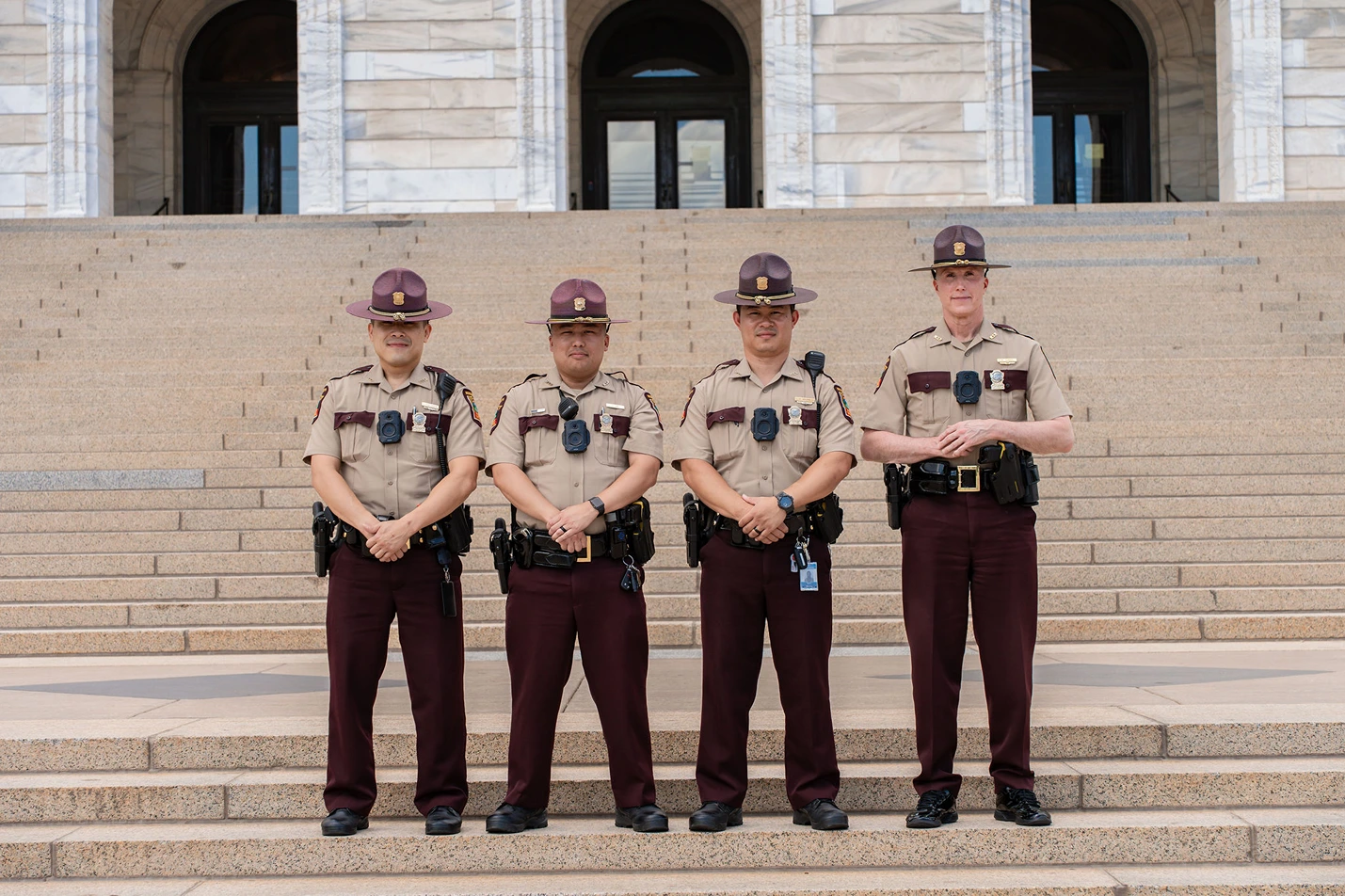 Four uniformed police officers standing in a row on stone steps in front of a building with large arched doorways.
