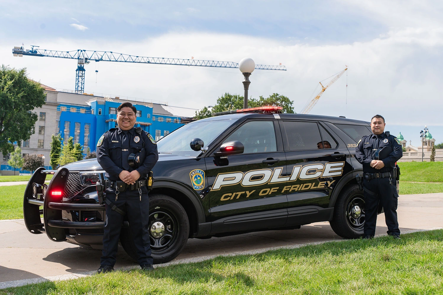 Two police officers standing beside a black City of Fridley police SUV with red lights on, outdoors in a park area.