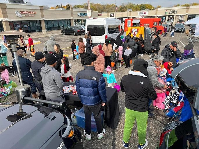 Community gathering at an outdoor trunk-or-treat event with families and children in costumes lined up near decorated vehicles and tables with candy.