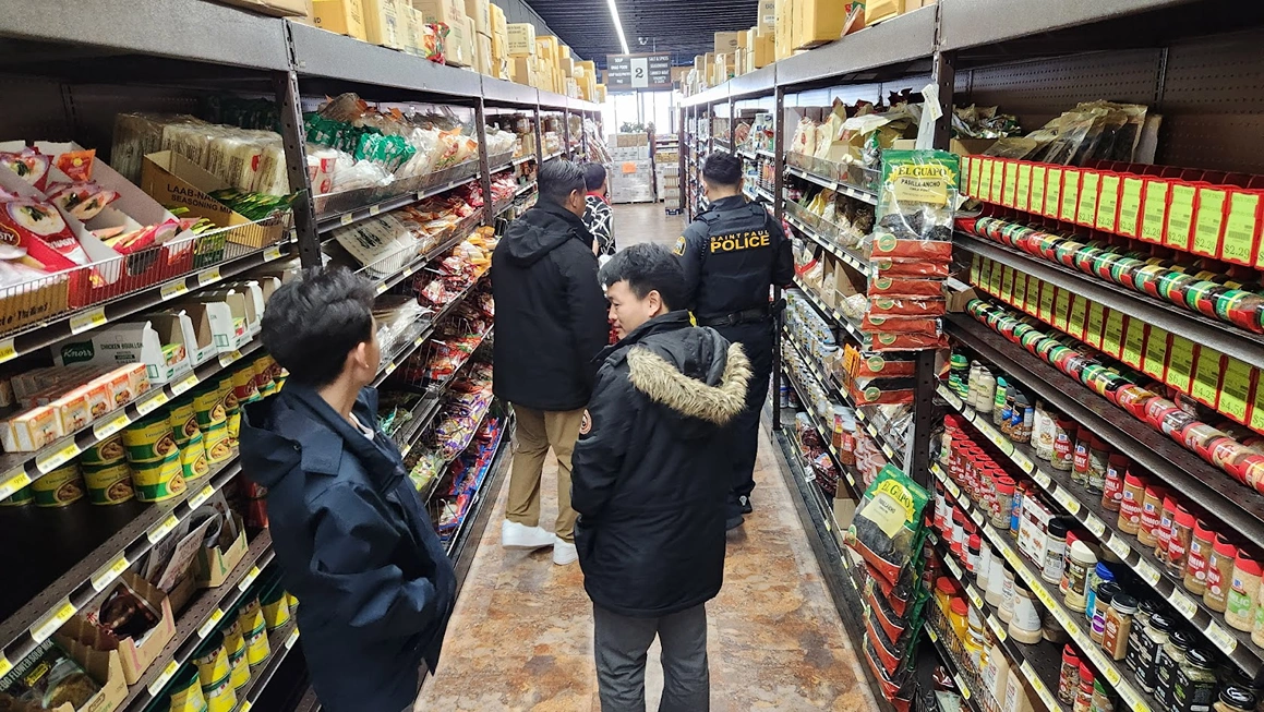 Four people, including a police officer, walking down an aisle of a grocery store with shelves stocked with various food products.