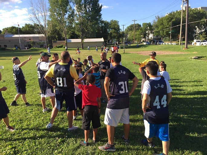 Group of youth and adult coaches gathered on a grassy field during a sunny day, some wearing numbered sports jerseys.