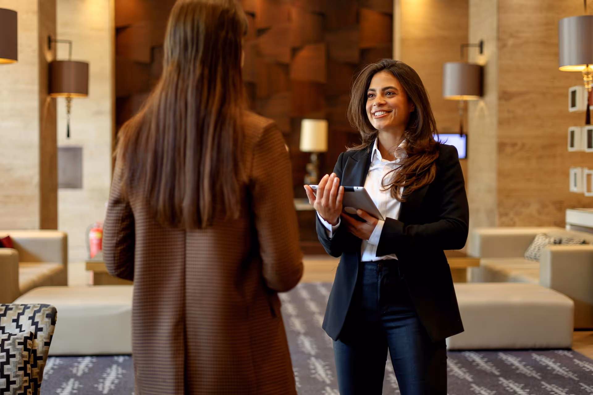 Two women meet indoors; one holds a tablet and smiles while talking to the other in a business setting.