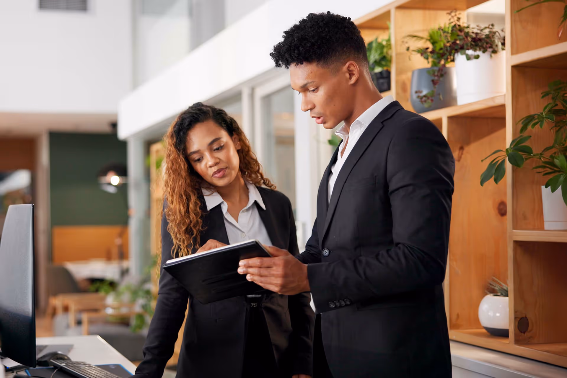 Two professionals in suits stand in an office, reviewing a document together near a shelf with potted plants.