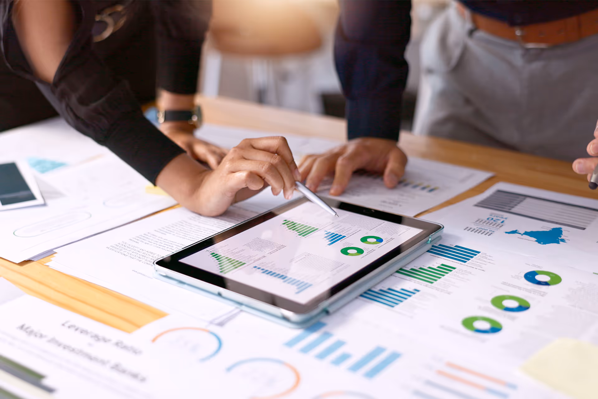 Hands pointing at charts on a tablet, surrounded by printed graphs and financial documents on a desk.