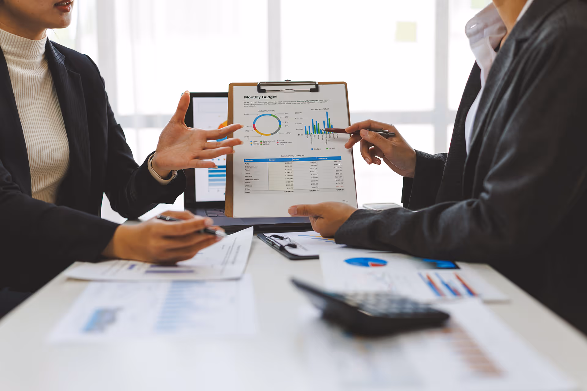 Two people in business attire review charts and graphs on paper and a clipboard during a meeting at a desk.