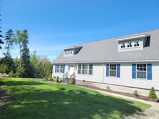 White house with gray roof and blue window shutters, surrounded by green lawn and trees under a clear blue sky.