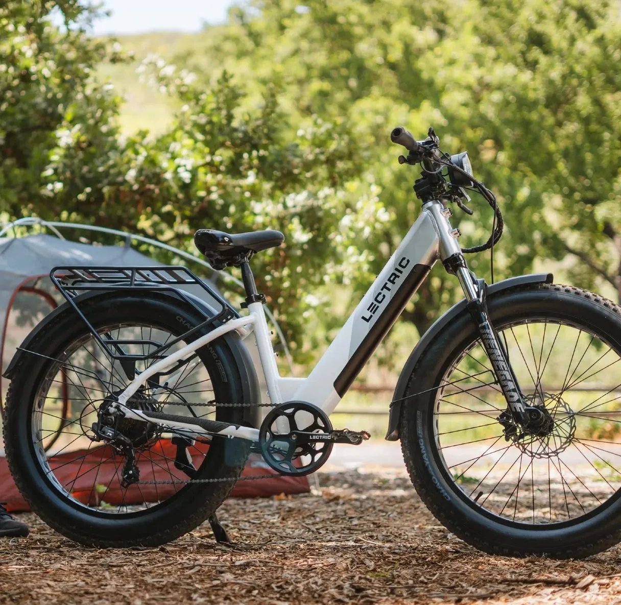 White Lectric electric bike with fat tires parked on wood chips in a green outdoor setting near a tent.