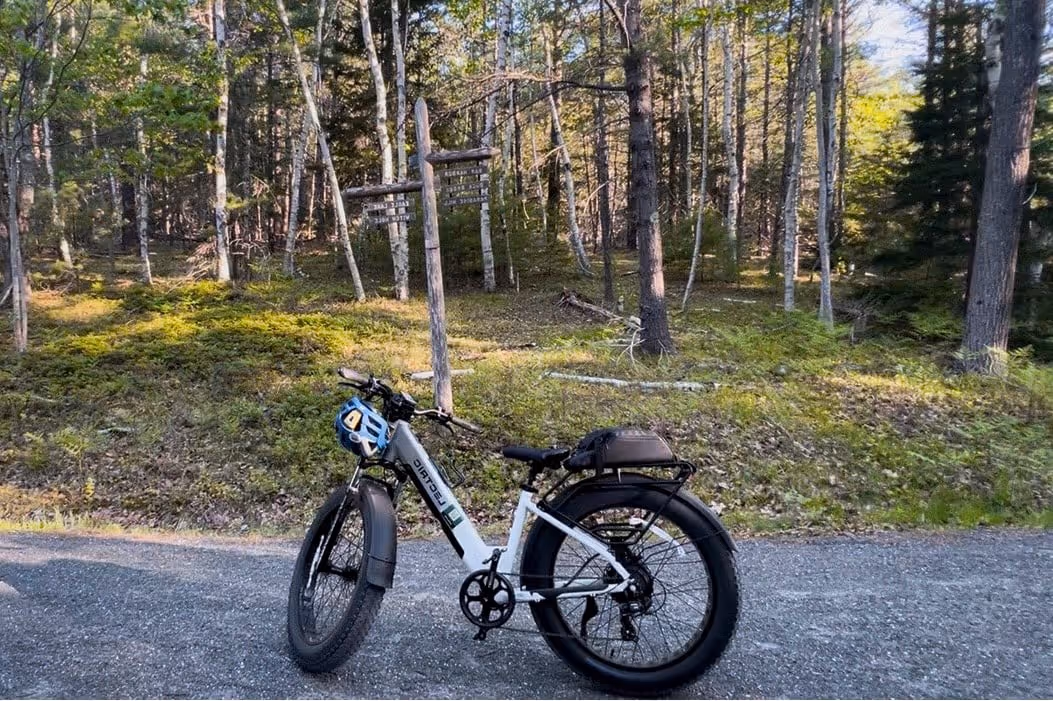 White electric fat-tire bike parked on a gravel road with a blue helmet hanging from the handlebars, in front of a forest with a wooden signpost.