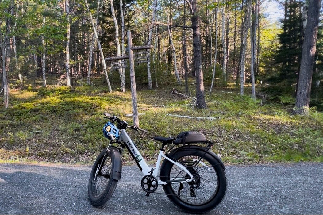 White electric fat-tire bike parked on a gravel road with a blue helmet hanging from the handlebars, in front of a forest with a wooden signpost.