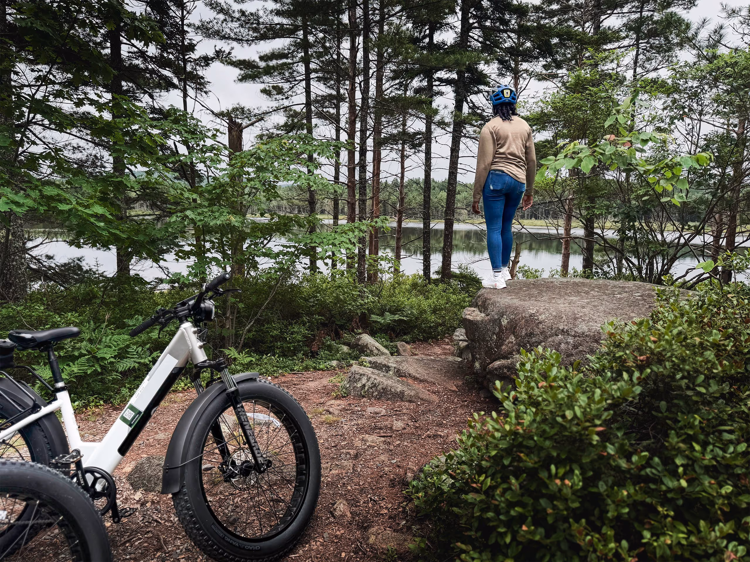 Person wearing blue jeans and a helmet standing on a large rock overlooking a forested lake, with an electric bike parked on a dirt trail nearby.
