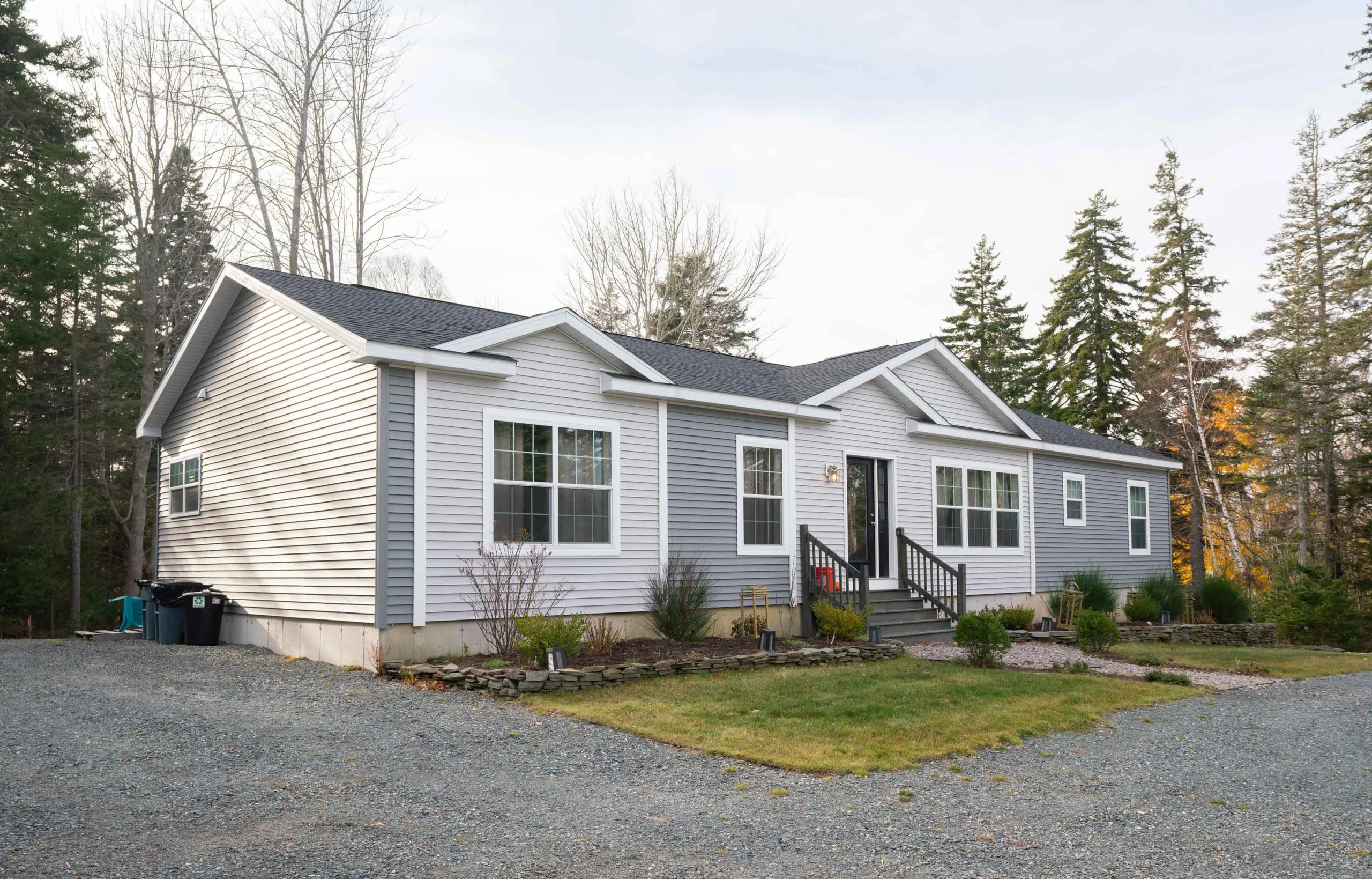 Single-story house with light gray siding, multiple windows, black front door, small porch stairs, surrounded by trees and a gravel driveway.