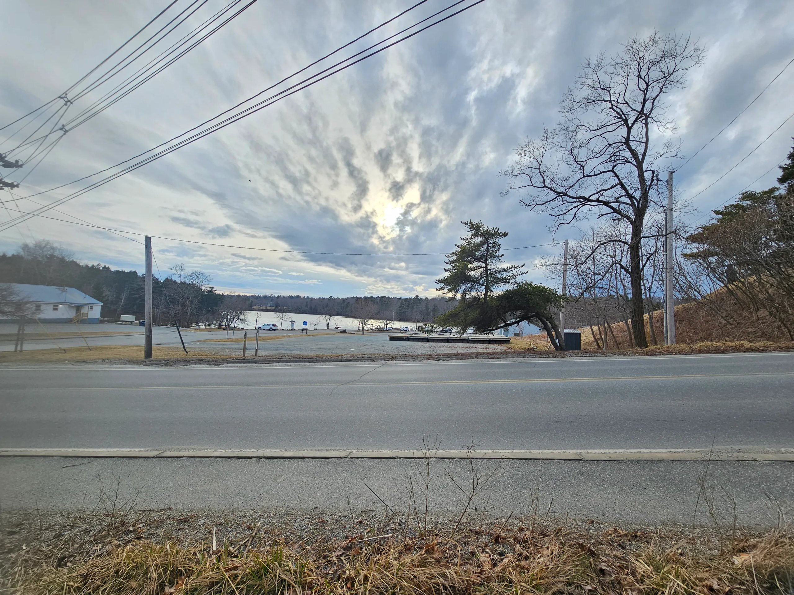 Overcast sky over a quiet lakeside area with a road, leafless trees, and a dock near the water.