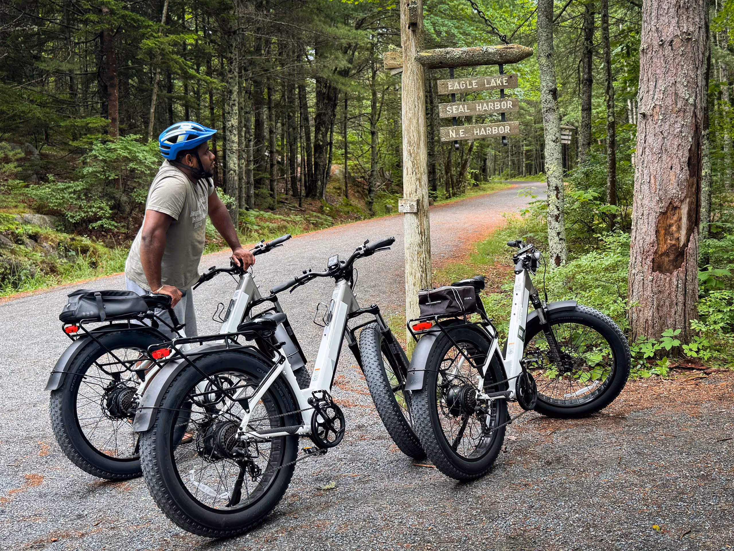 Man wearing a blue helmet standing next to three white electric bikes on a forest trail near a wooden signpost pointing to Eagle Lake, Seal Harbor, and N.E. Harbor.