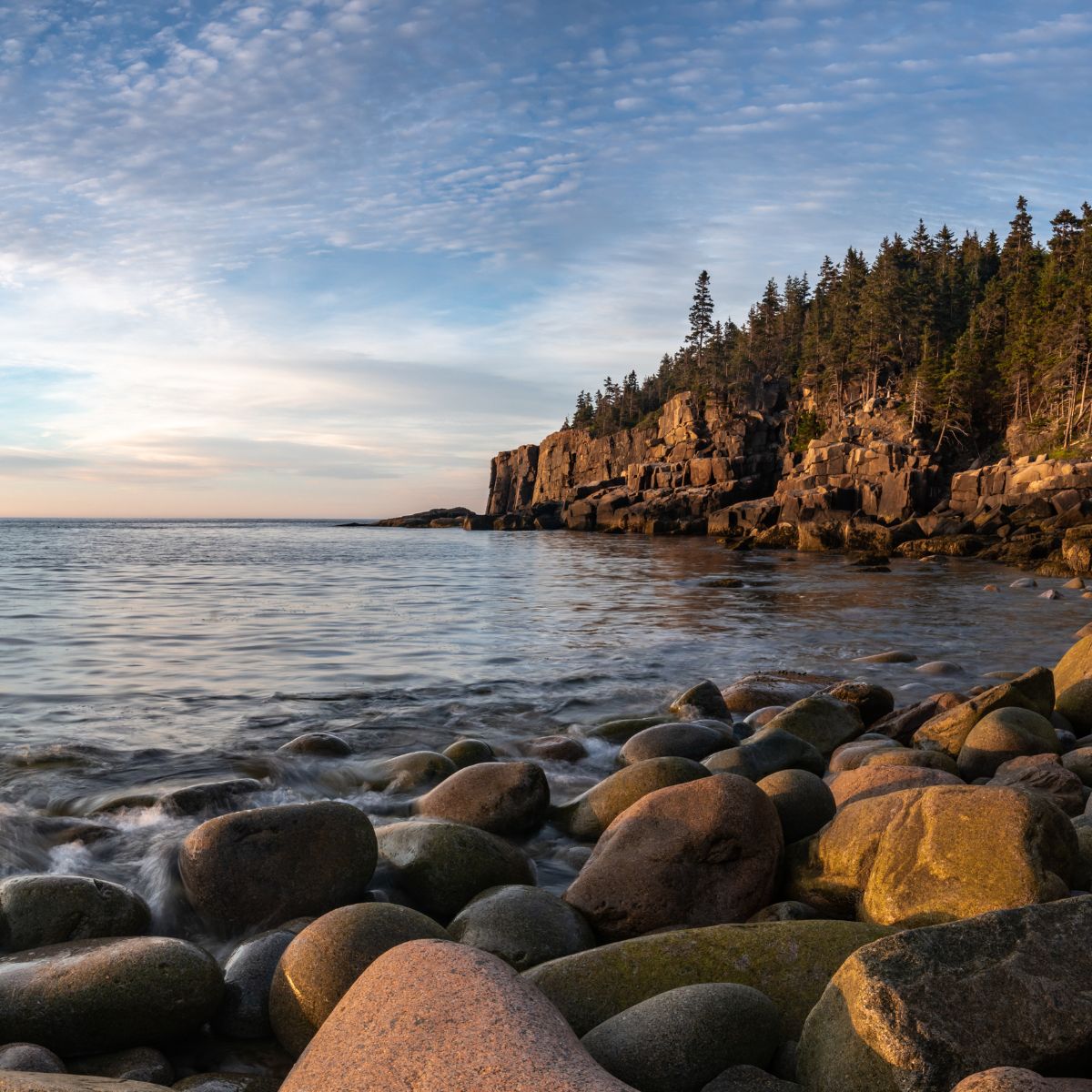 Rocky shoreline with large boulders, calm ocean water, and forested cliffs under a partly cloudy sky at sunset.